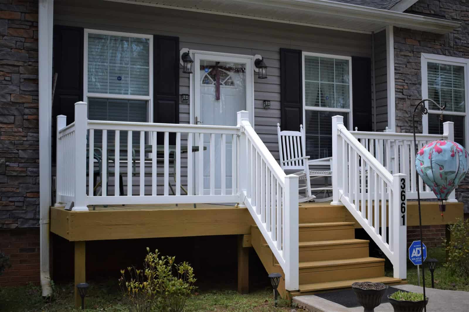 A house front porch with white railings, light-colored wooden steps, and two white rocking chairs. The exterior includes gray siding, stone accents, black shutters, and a white front door with glass panels. A colorful hot air balloon decoration and ADT security sign are visible.