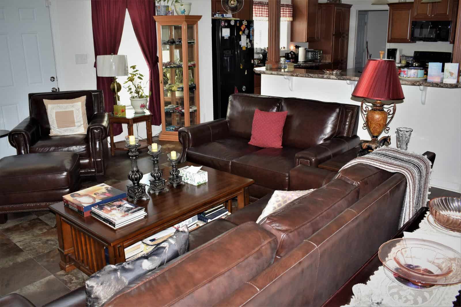 A cozy living room with dark brown leather sofas and armchairs arranged around a wooden coffee table. The table is adorned with candles, magazines, and decor items. The room features a kitchen area with a bar counter and stools in the background.
