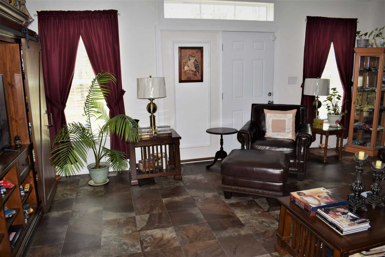 A cozy living room with brown leather armchair, side table, and lamp. Red curtains frame two windows on either side of a white front door. Shelves display various knick-knacks. A potted plant is in the corner. Magazines are on a coffee table in the foreground.