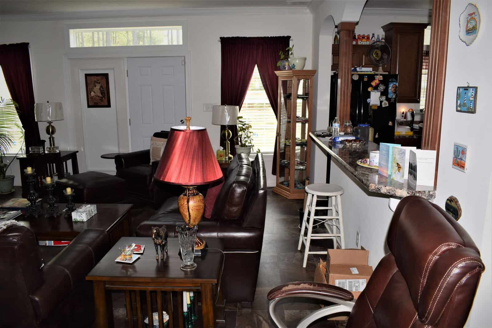 A cozy living room with brown leather furniture and a red lamp. The room has dark red curtains, a mix of wooden and glass furniture, and various decorative items. A breakfast bar with stools separates the kitchen. The kitchen has a black refrigerator and various items on the counter.