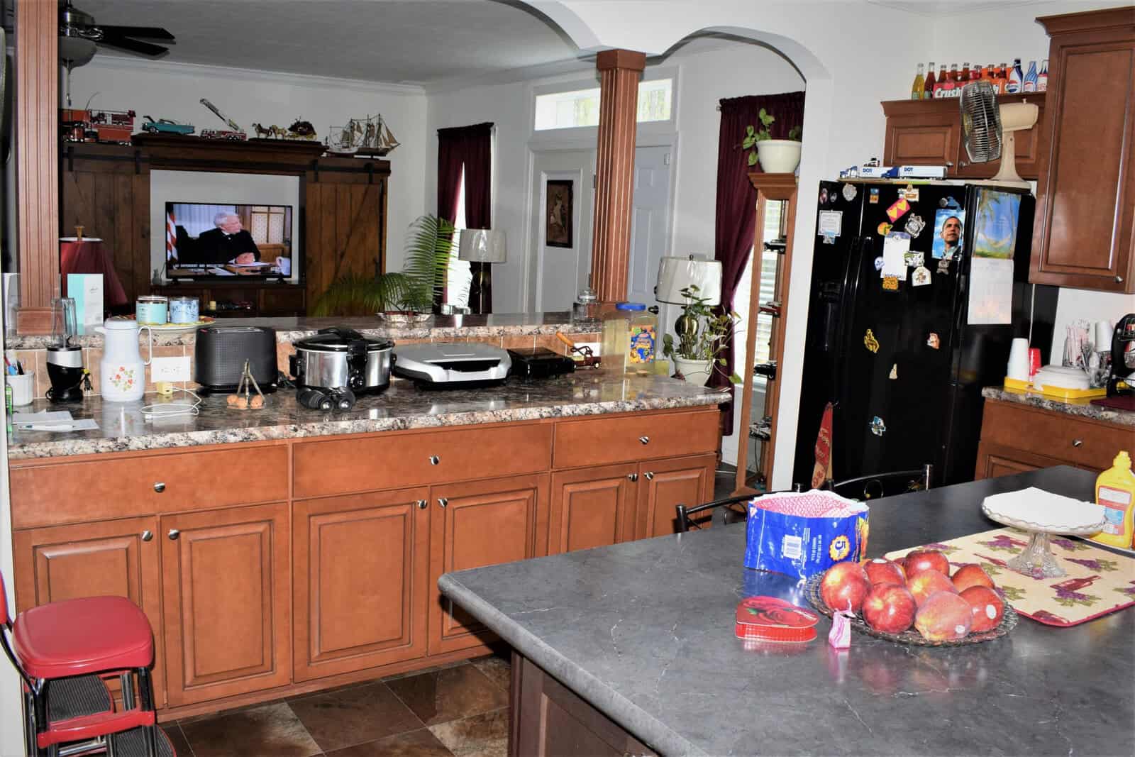A kitchen with brown cabinets, a dark countertop, and a black refrigerator covered with magnets. The counter has apples, soap, a bag of bread, various appliances, and a bottle of cooking oil. A TV in the background shows a person speaking.