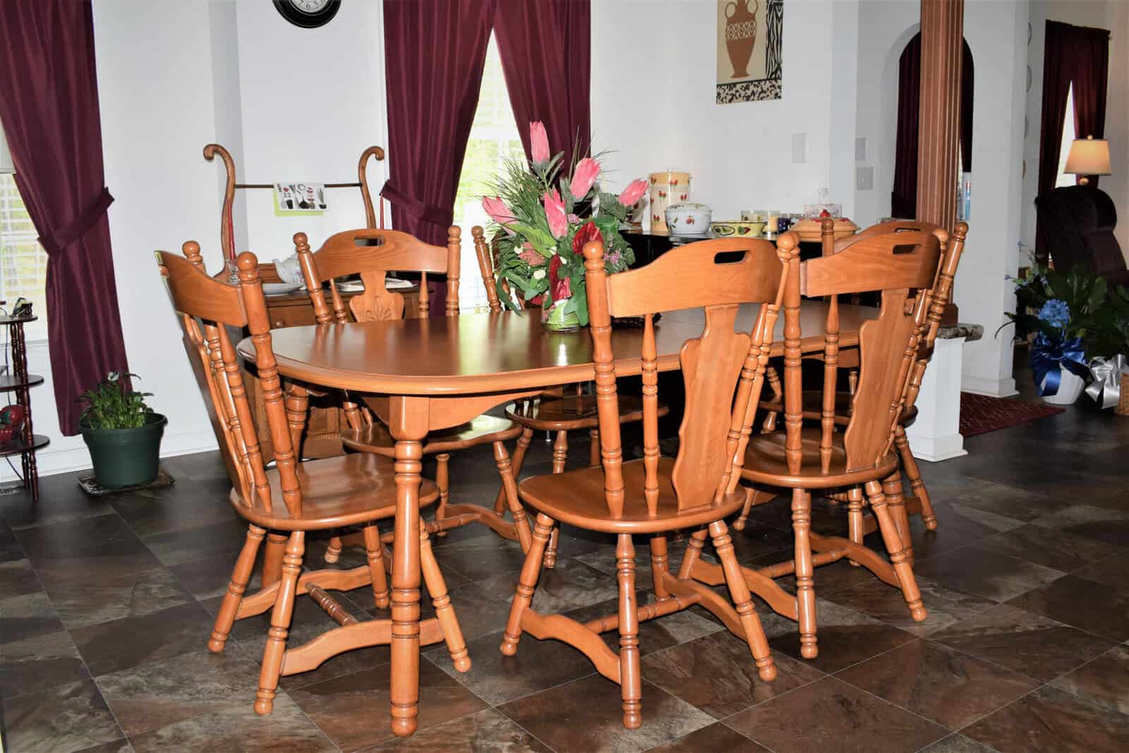 A wooden dining table with six matching chairs is set in a room with tiled flooring and deep burgundy curtains. A floral arrangement decorates the table. The background includes various household items, potted plants, and artwork on the walls.