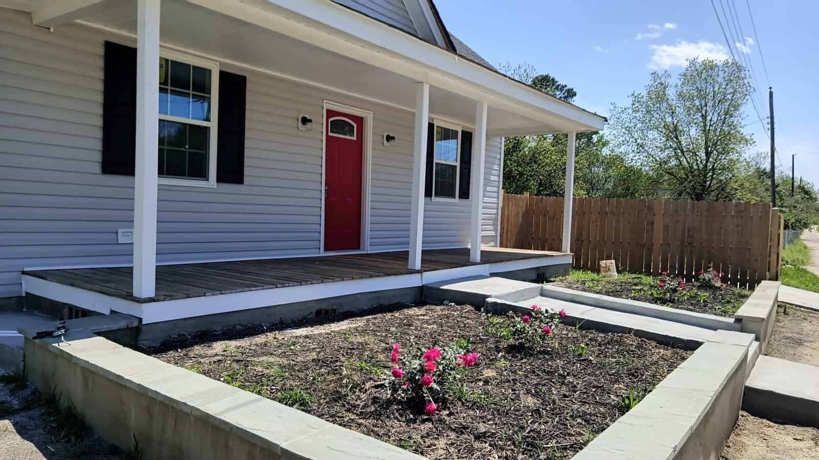 A single-story house with gray siding and a red front door, featuring a wooden porch and garden beds with blooming pink flowers in the foreground.