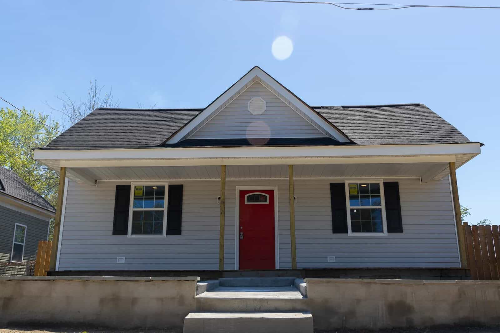 A symmetric two-story house with a red front door and gray siding under a clear blue sky, exhibiting lens flare effects from the sun.
