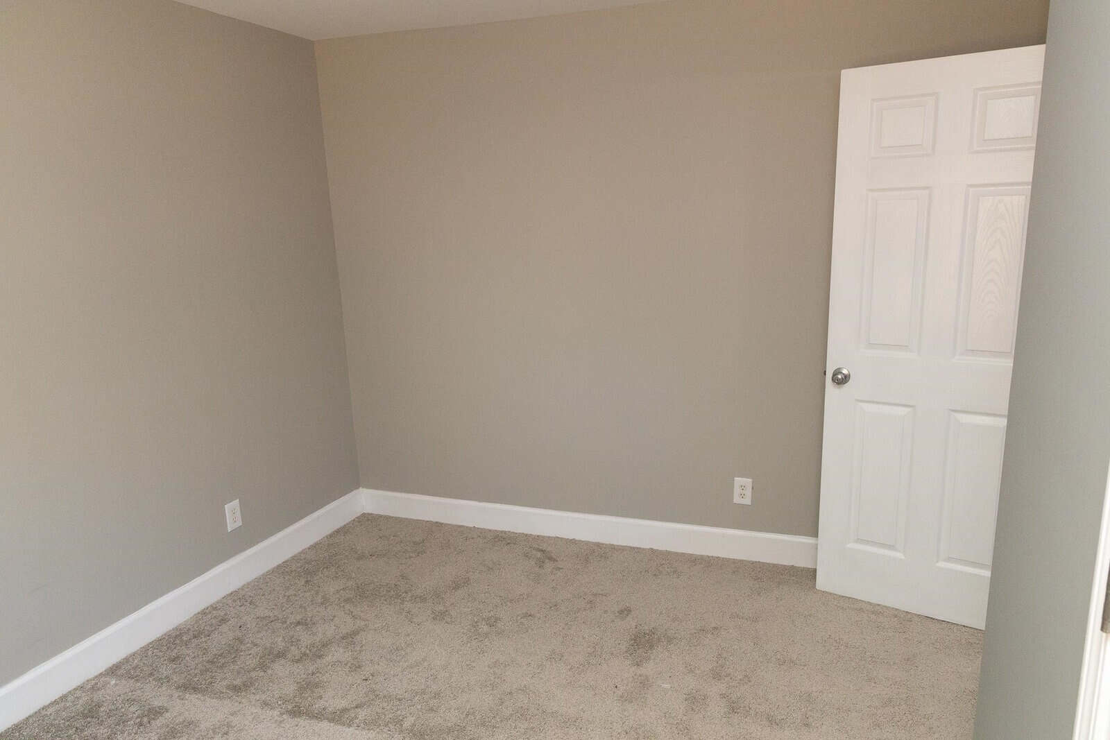 An empty room with beige walls and carpet flooring featuring a closed white door.