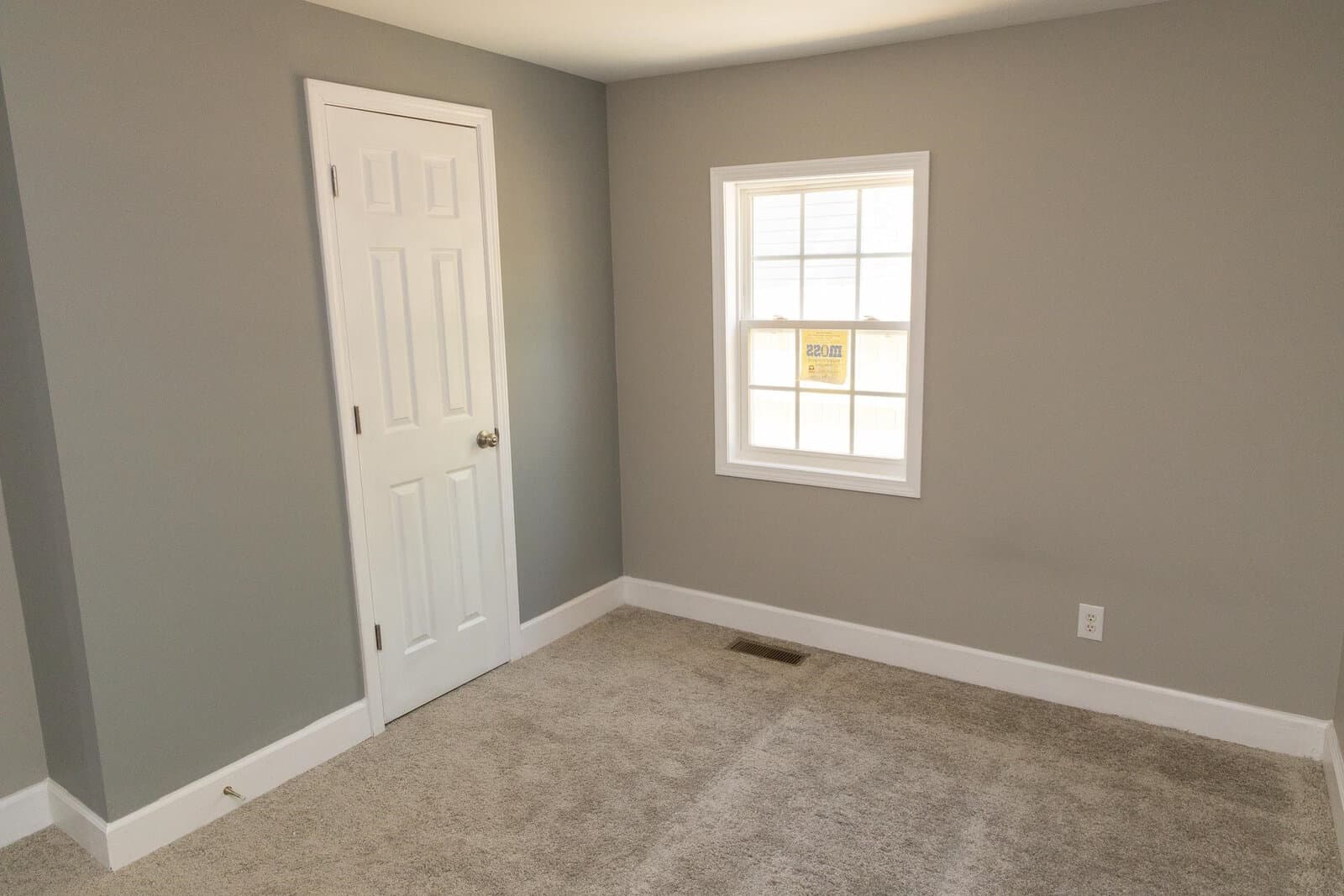A vacant room with gray walls, a white door, and a window with a view of the exterior, featuring beige carpeting.