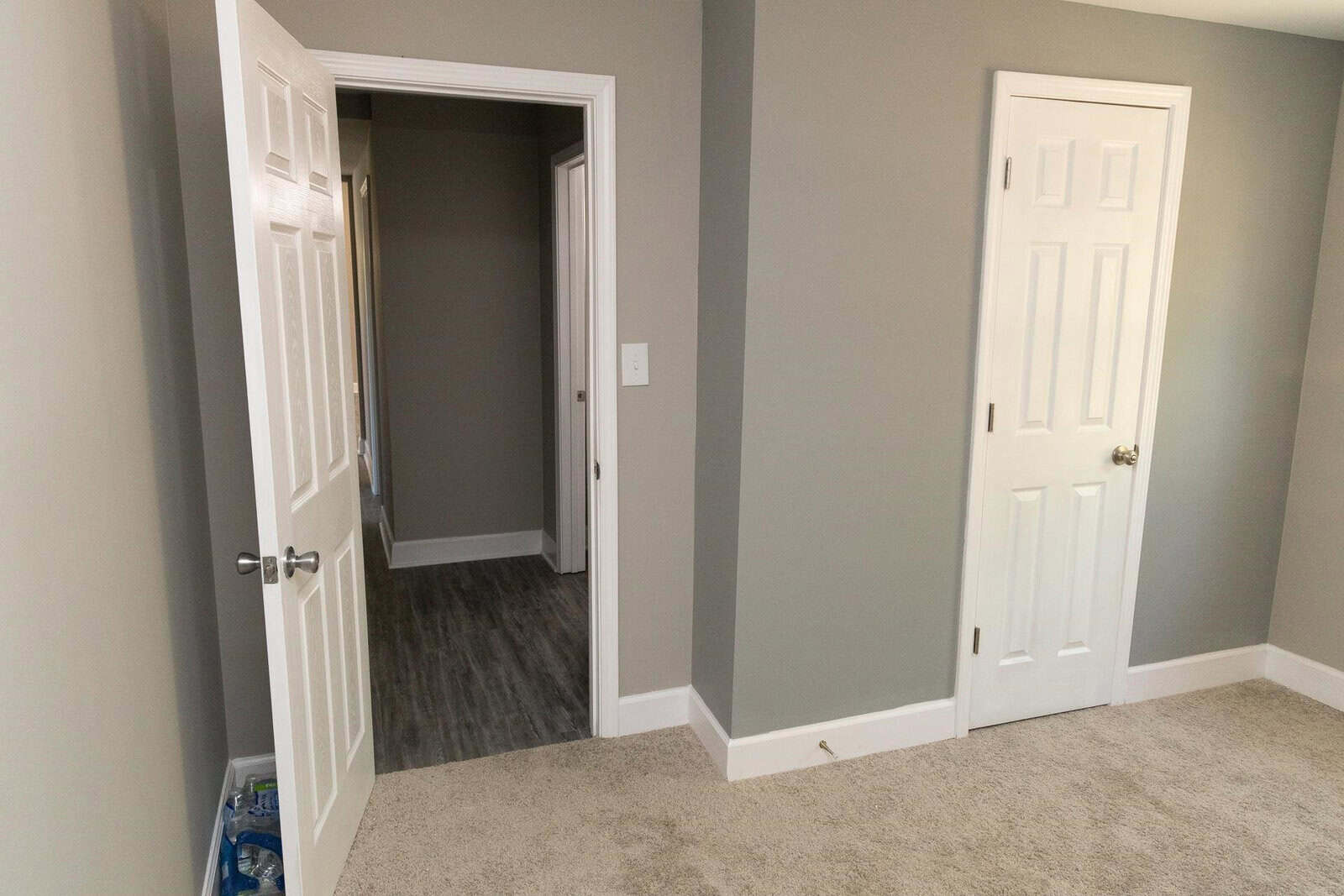 A residential interior featuring an open door leading to a hallway with hardwood flooring, adjacent to a closed door, both set in walls painted in a neutral gray color, complemented by beige carpeting in the foreground.