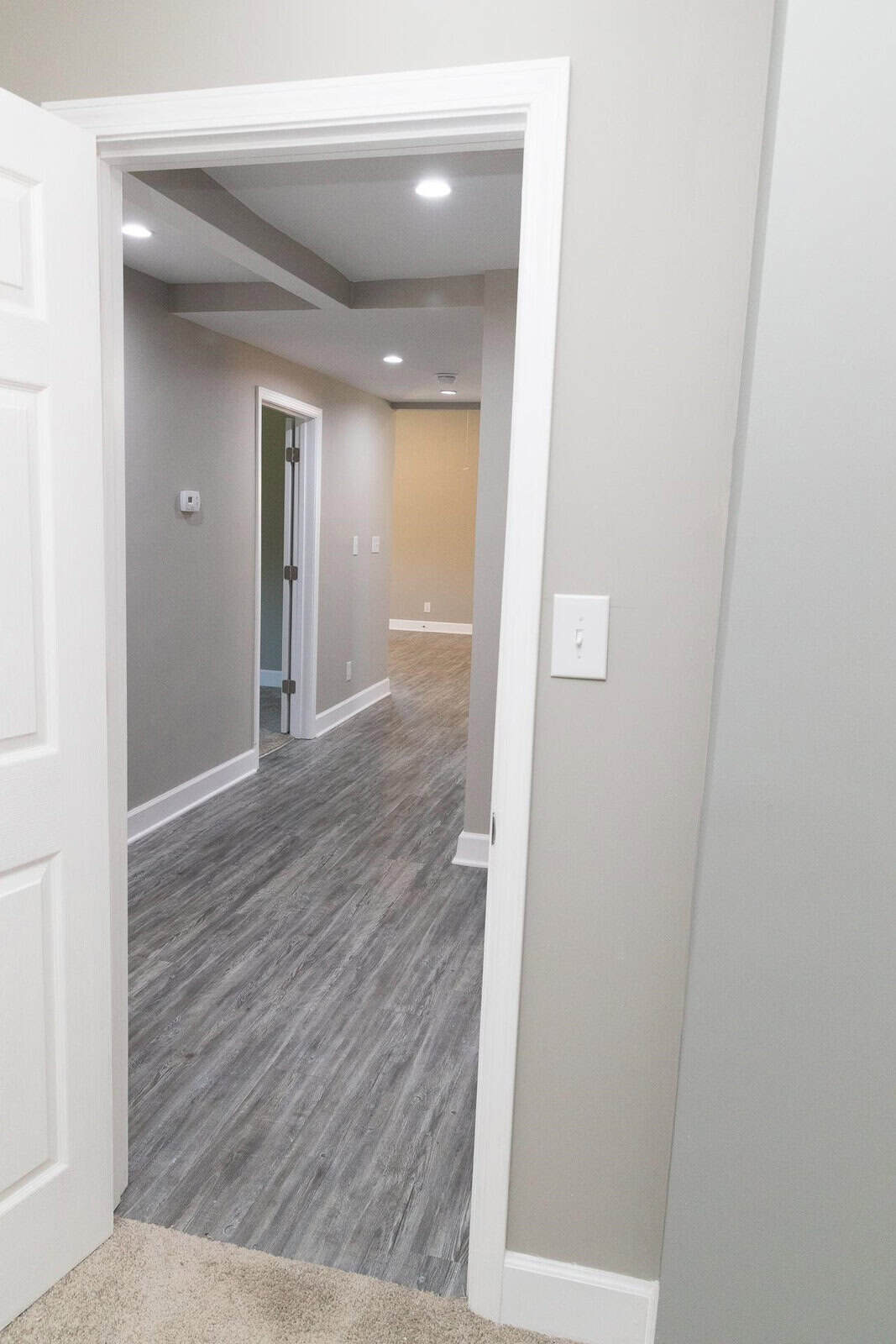 A view through an open doorway showing an empty hallway with gray wood patterned flooring, white walls, and ceiling recessed lighting.