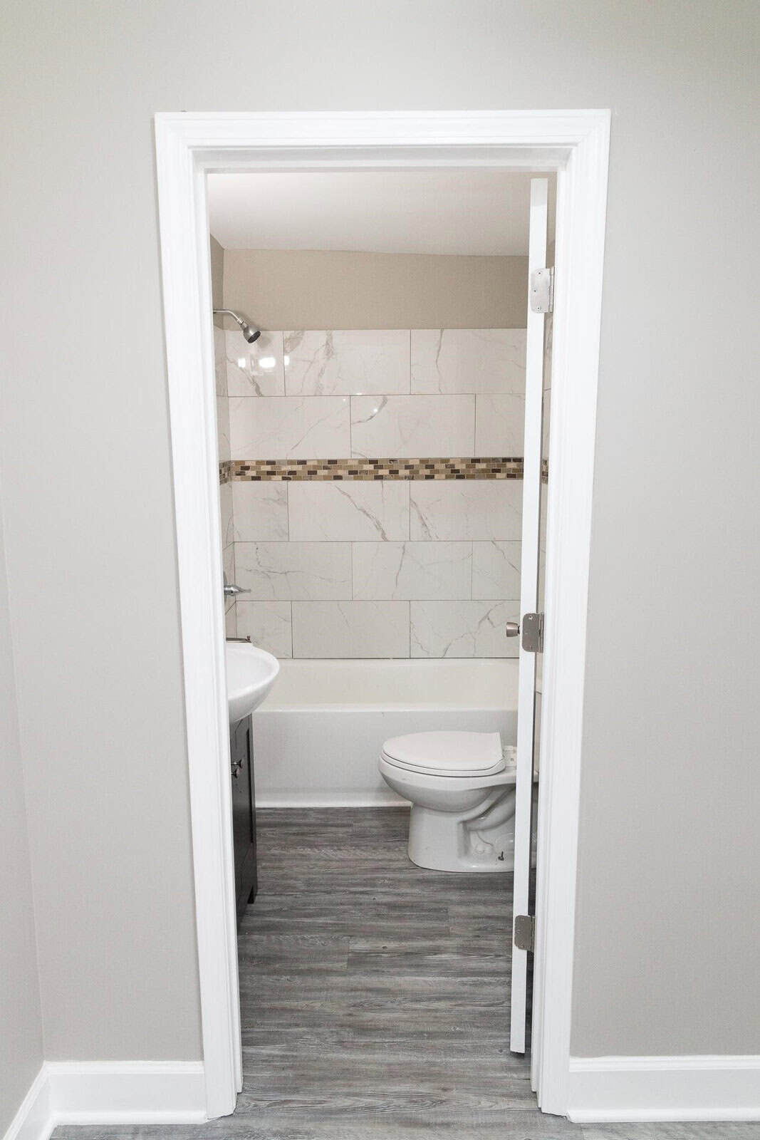 A modern bathroom viewed from the doorway, featuring a white toilet and sink, with light gray flooring and white marble-like tiles on the walls, accented by a decorative mosaic tile strip.