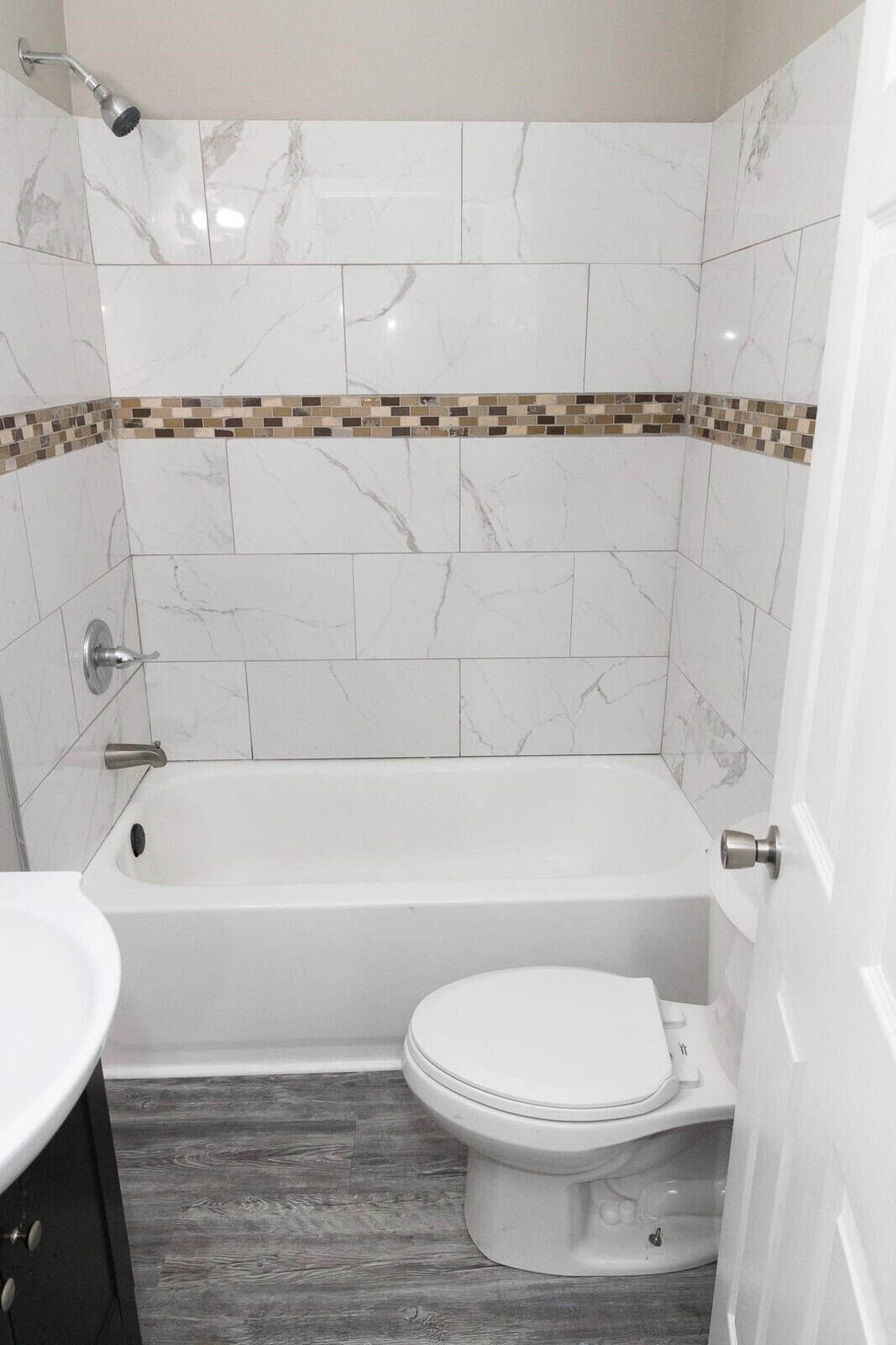 A modern bathroom with white marble-like tiles, a small accent tile border, a white bathtub, a toilet, and gray flooring.