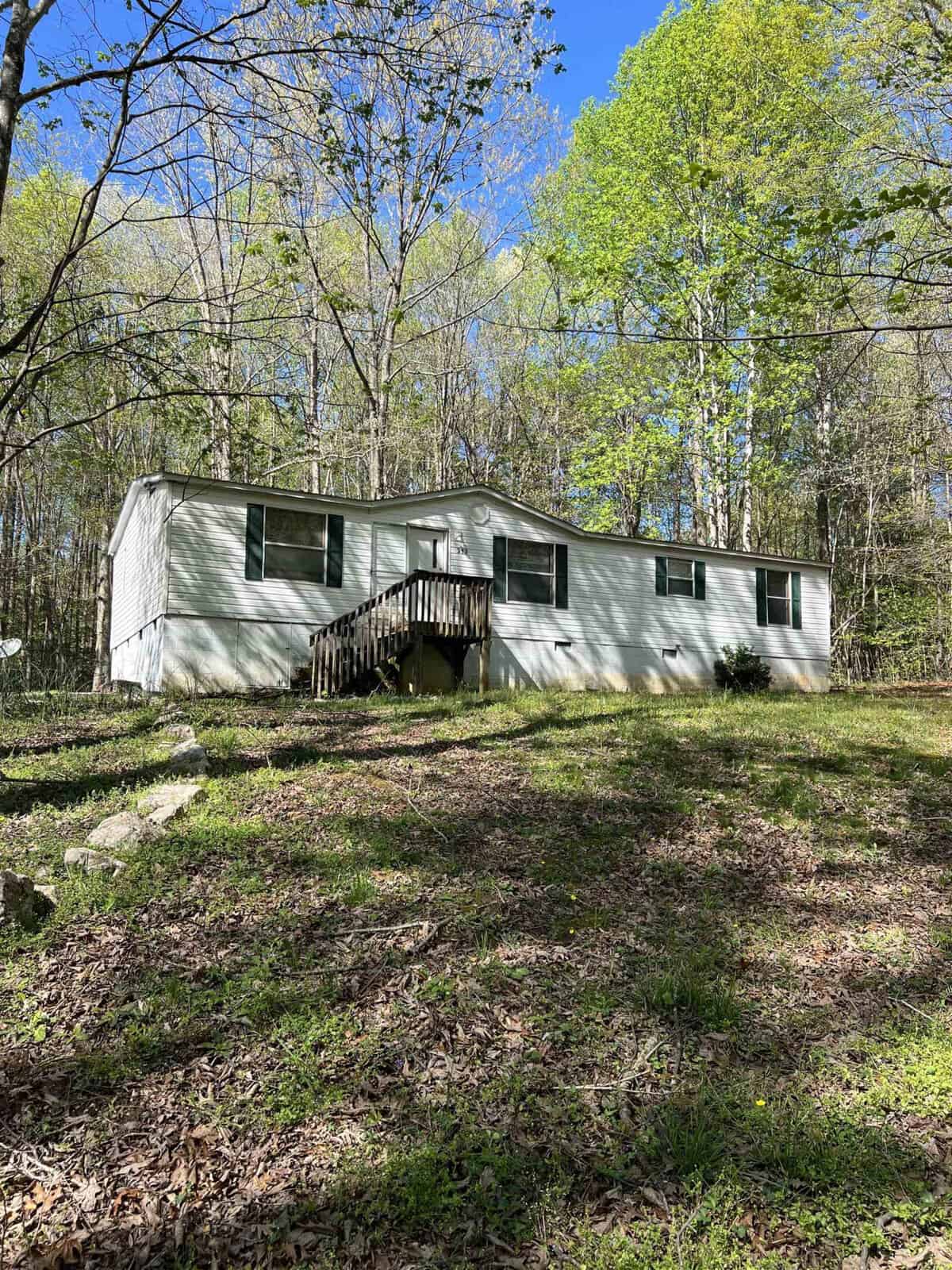 A single-story mobile home with a weathered wooden staircase situated in a wooded area during daytime.