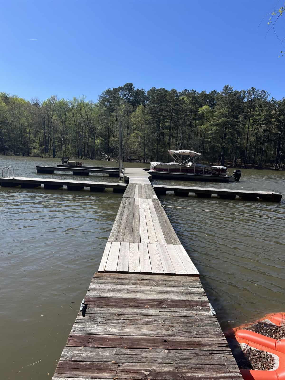 A wooden pier stretches out into a body of water leading to a dock where a pontoon boat is moored on a sunny day with a forested shoreline in the background.