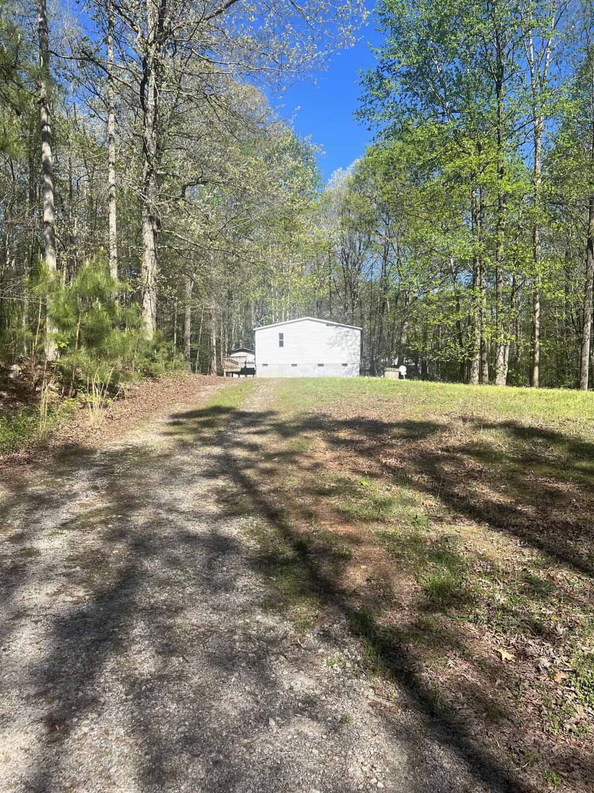 A small white building surrounded by trees on a sunny day, with tree shadows cast onto a grassy area and a dirt pathway.