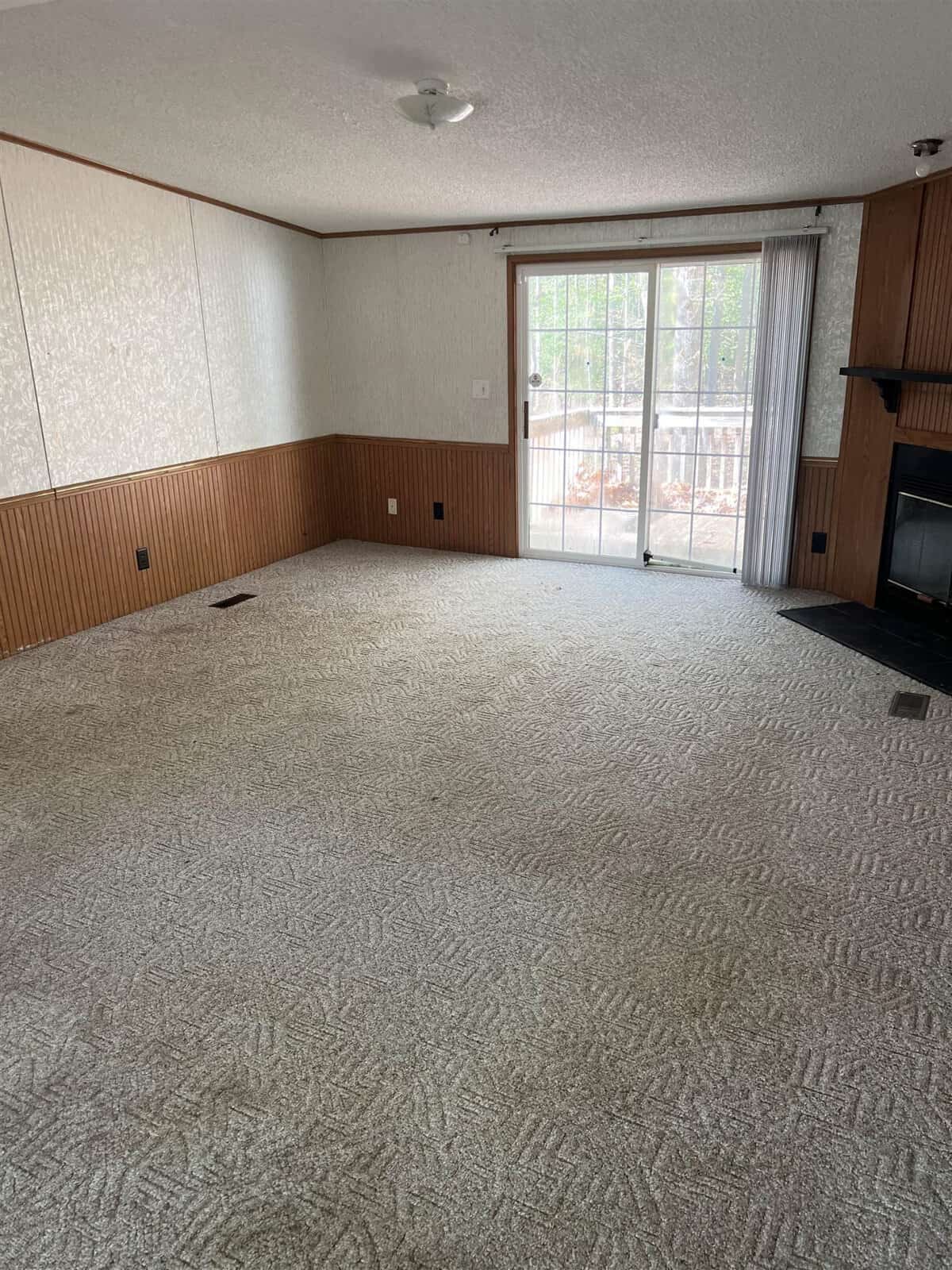 An empty room with beige walls, a section with wood paneling, carpeted flooring, and a fireplace in the corner near a large window with a view of a fence outside.