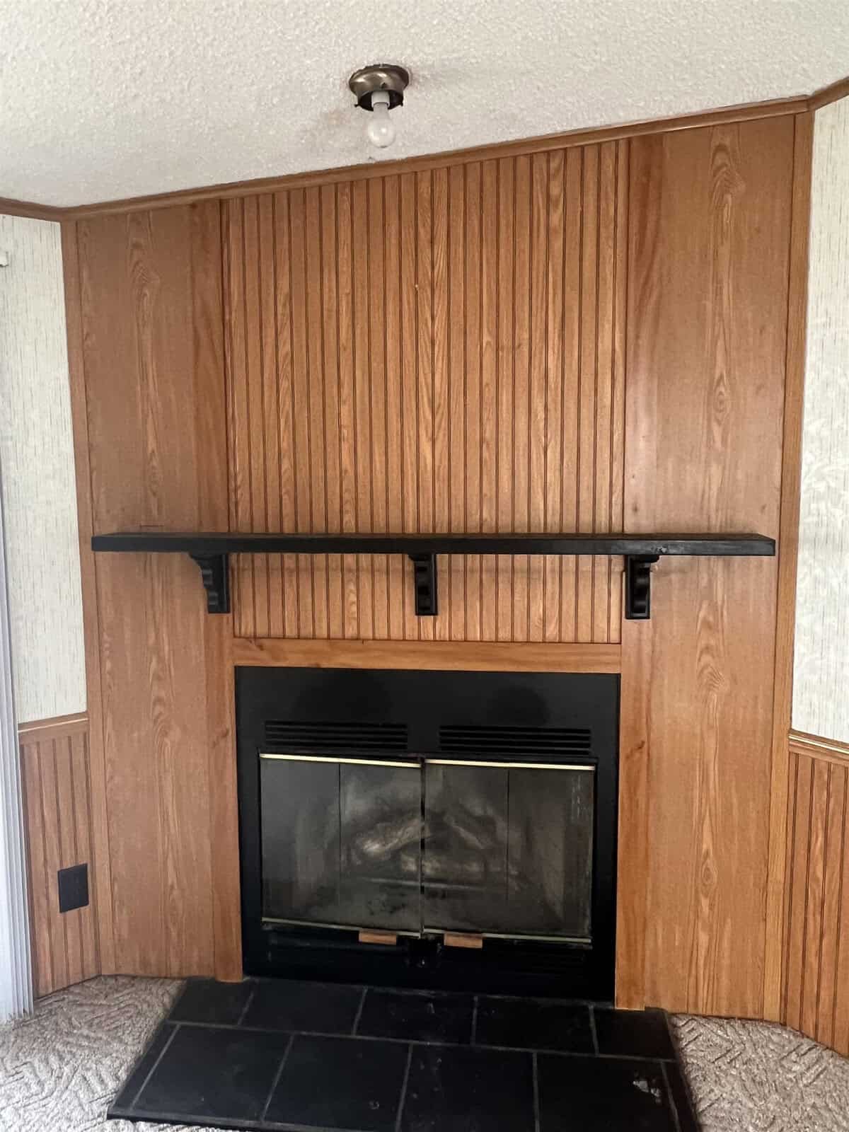 A wood-paneled room with a black fireplace and mantle, under a simple ceiling light fixture.