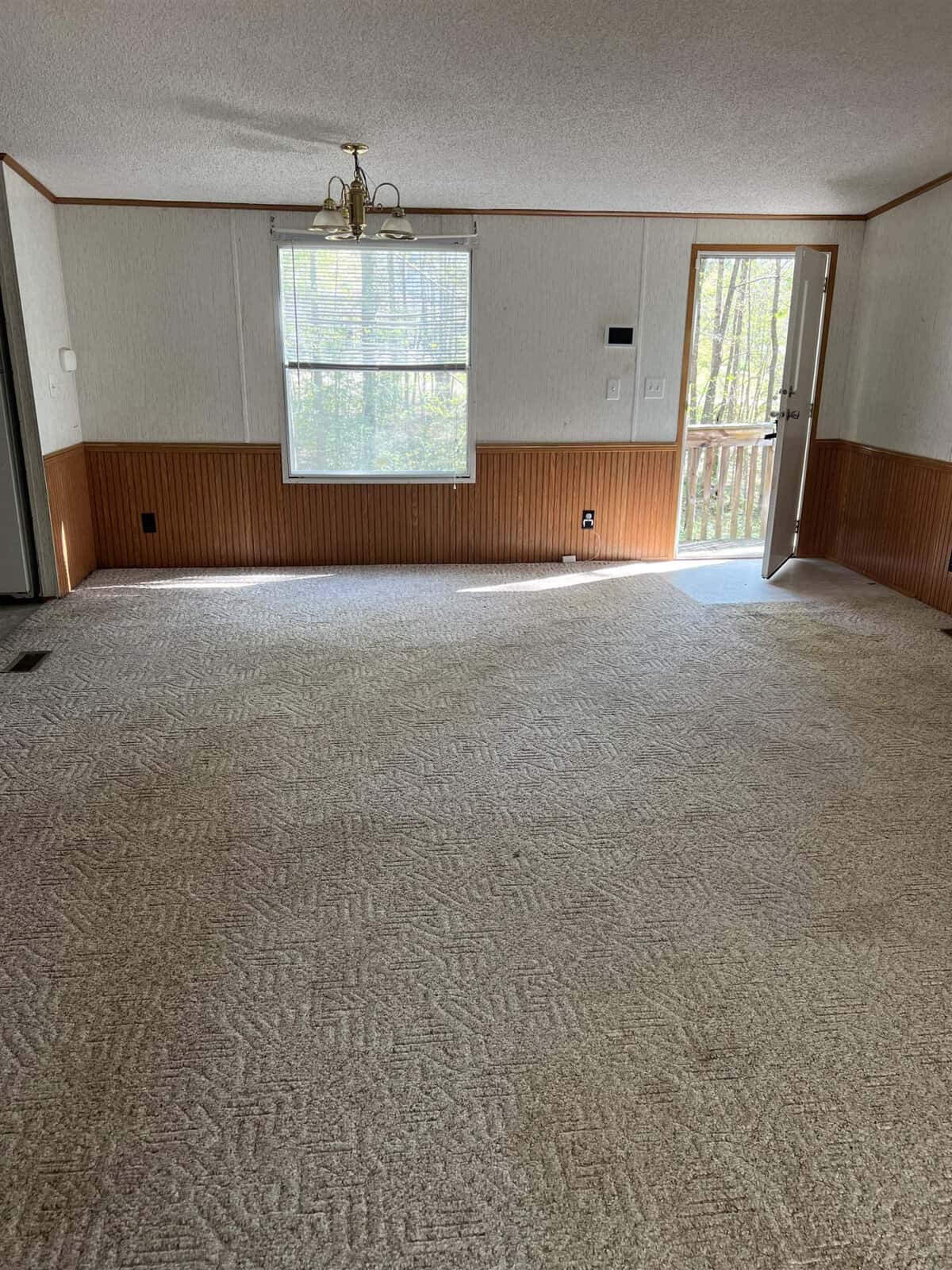 An empty room featuring beige carpeting, white walls with wooden wainscoting, two windows providing natural light, and a chandelier hanging from the ceiling.