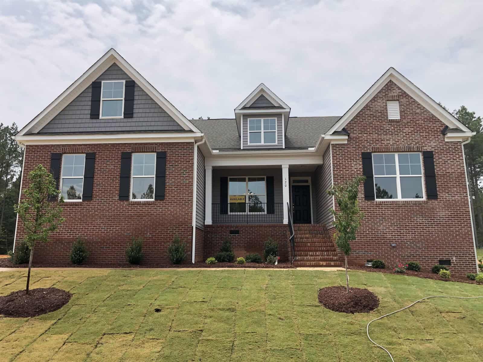 A newly built brick house with black shutters, three front gables, and a covered porch. The front yard has fresh sod, young trees, and small shrubs, with a hose lying on the grass.