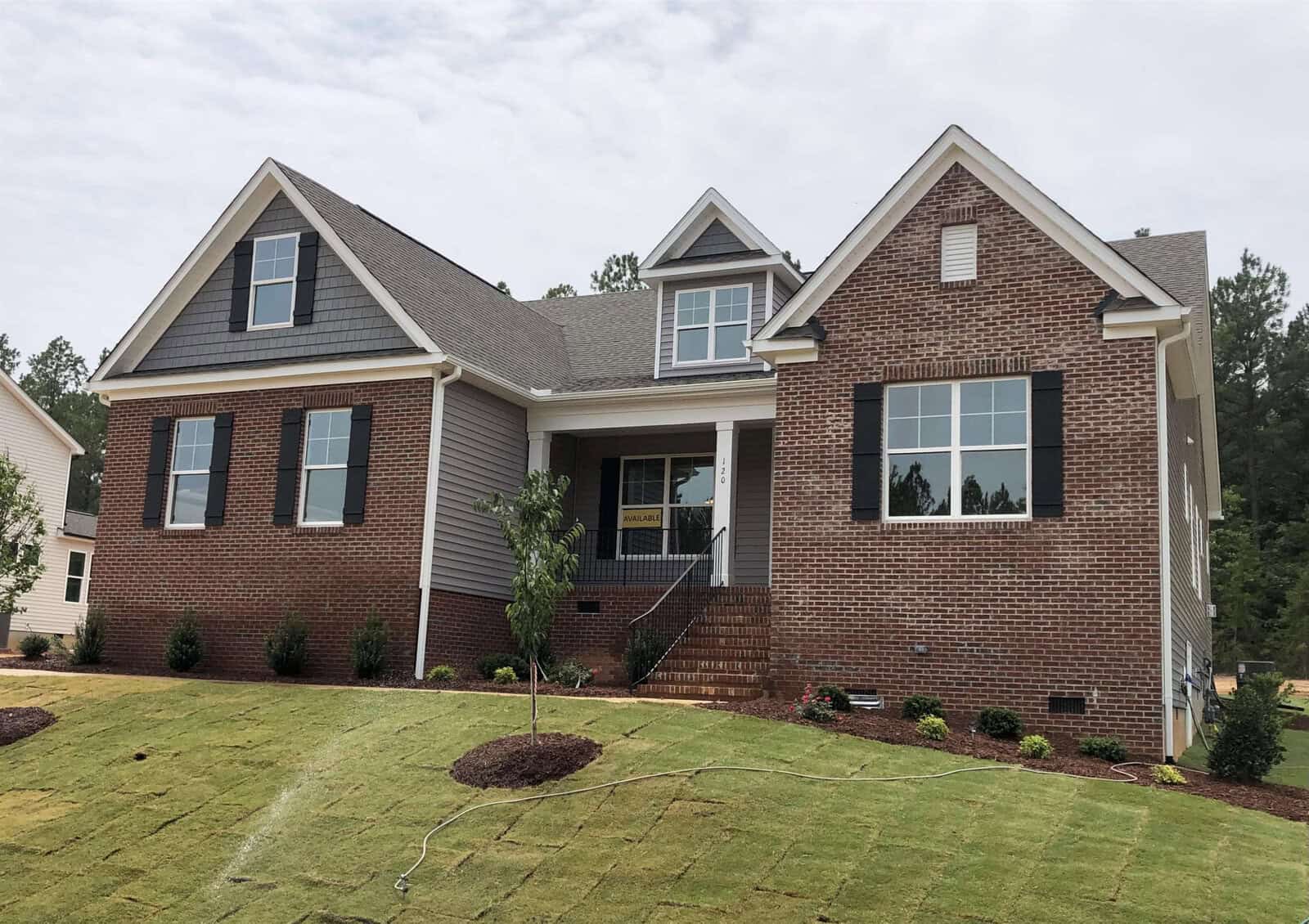 A newly built, single-story brick and siding house with a front porch, dark shutters, and a freshly landscaped lawn. A small tree and shrubs are planted across the yard under a cloudy sky.