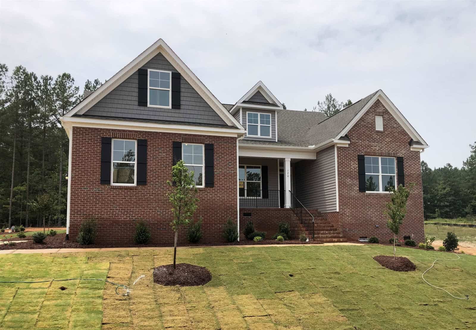 A newly built brick house with black shutters and gray siding, situated on a freshly sodded lawn with young trees and shrubs. The home features a small front porch and is set against a backdrop of trees and a partly cloudy sky.