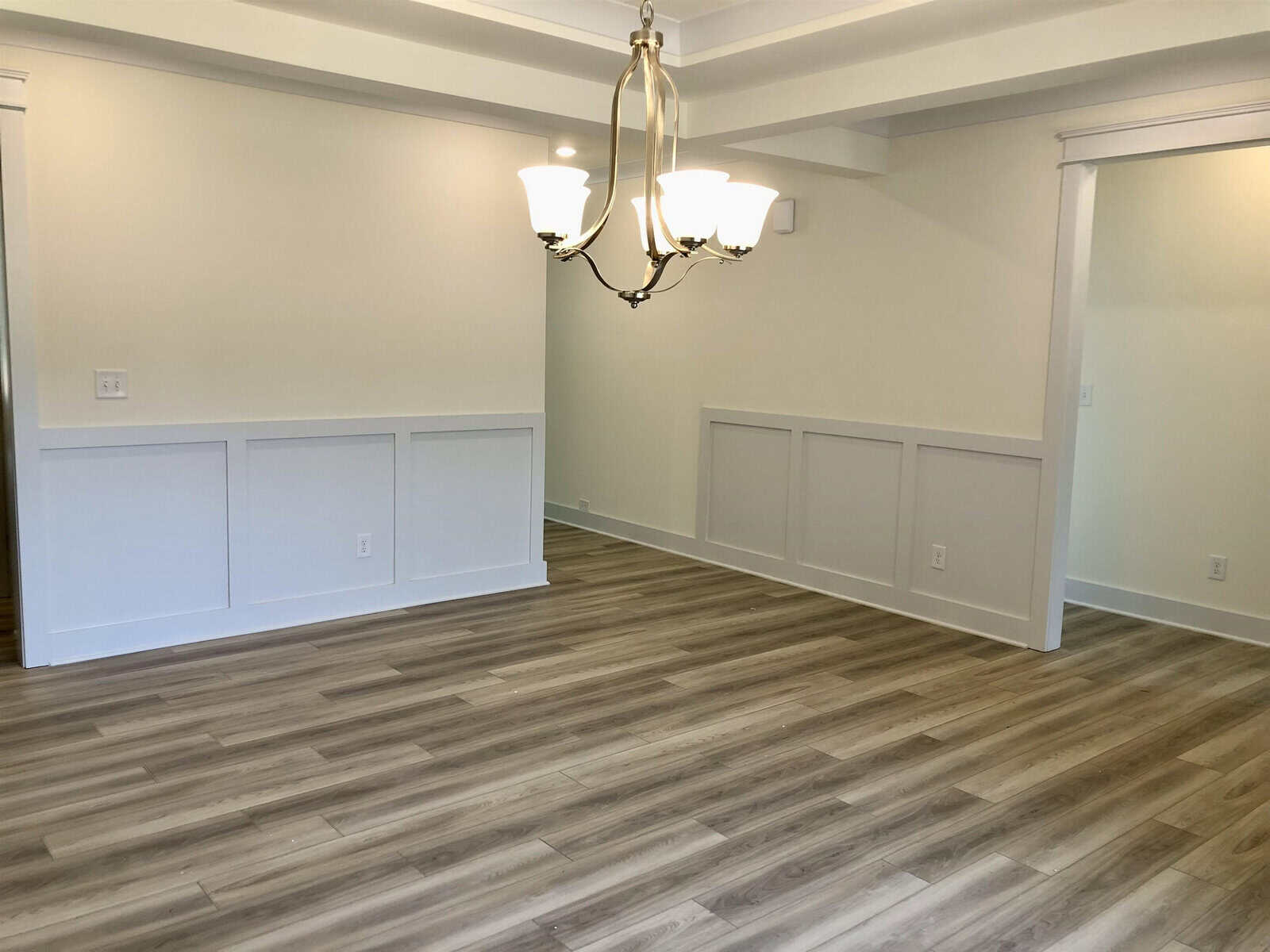 A bright, empty room with light wood flooring, cream-colored walls, white wainscoting, a tray ceiling, and a modern chandelier hanging from the ceiling.