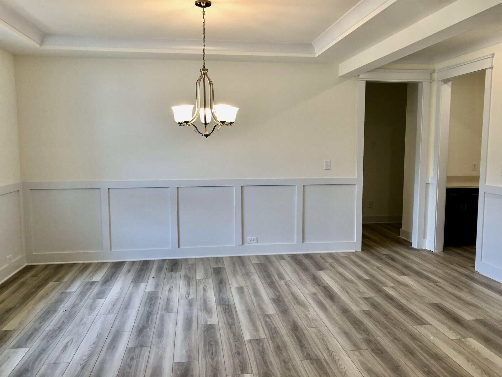 A bright, empty dining room with light wood flooring, white wainscoting, cream-colored walls, a modern chandelier, and an open doorway leading to another room.