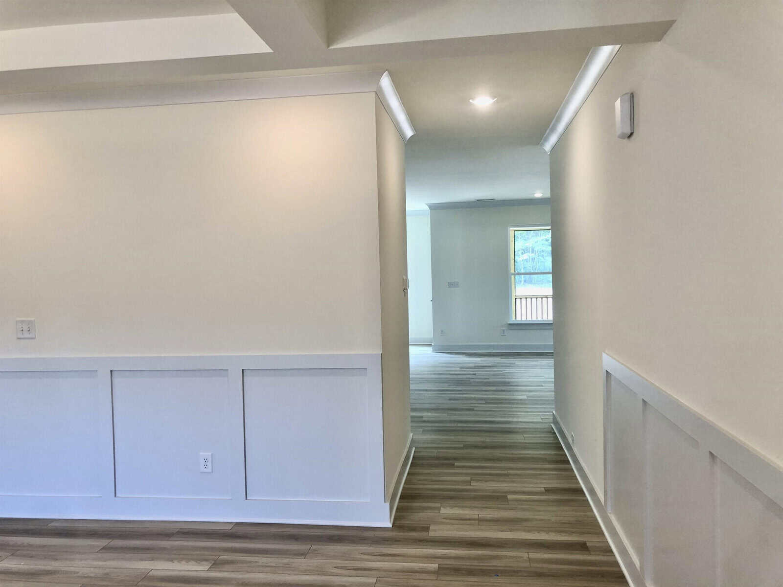 A hallway in a modern home with light-colored walls, white wainscoting, crown molding, and wood-style floors. Natural light streams through a window at the far end of the hallway.