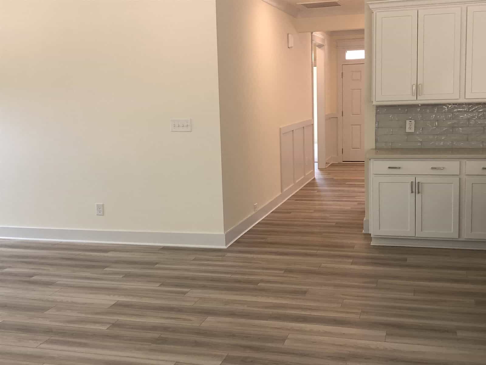 A bright, empty room with light wood flooring, cream-colored walls, white kitchen cabinets, and a textured backsplash. The image shows a hallway leading to a front door in the background.