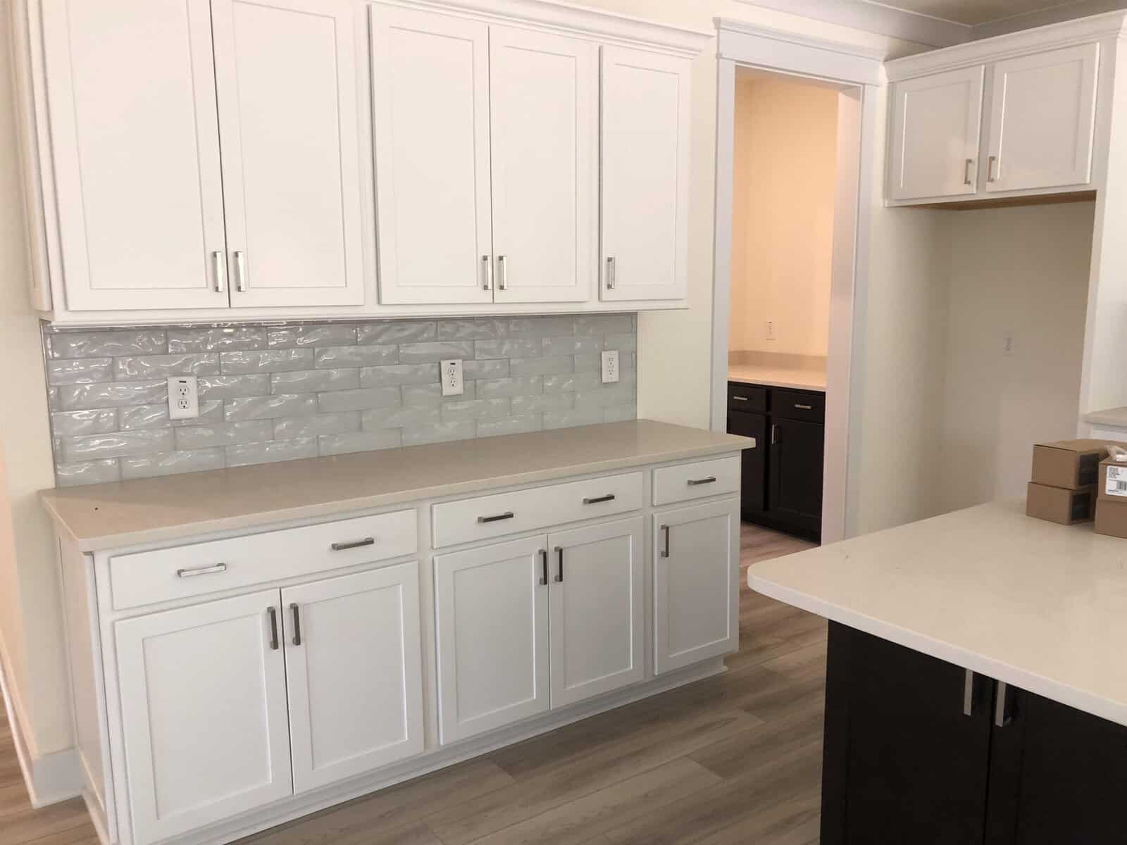 A modern kitchen with white cabinets, silver handles, light countertops, a light gray textured tile backsplash, and light wood flooring. An adjacent room with dark cabinets is visible through a doorway.