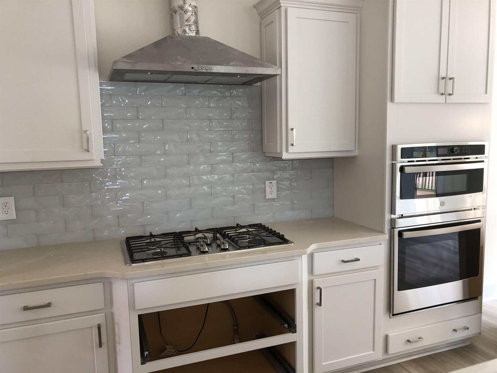 A modern kitchen features white cabinets, a stainless steel gas cooktop, a matching vent hood, and built-in double ovens. The area under the cooktop is open, with a glossy light gray subway tile backsplash behind.