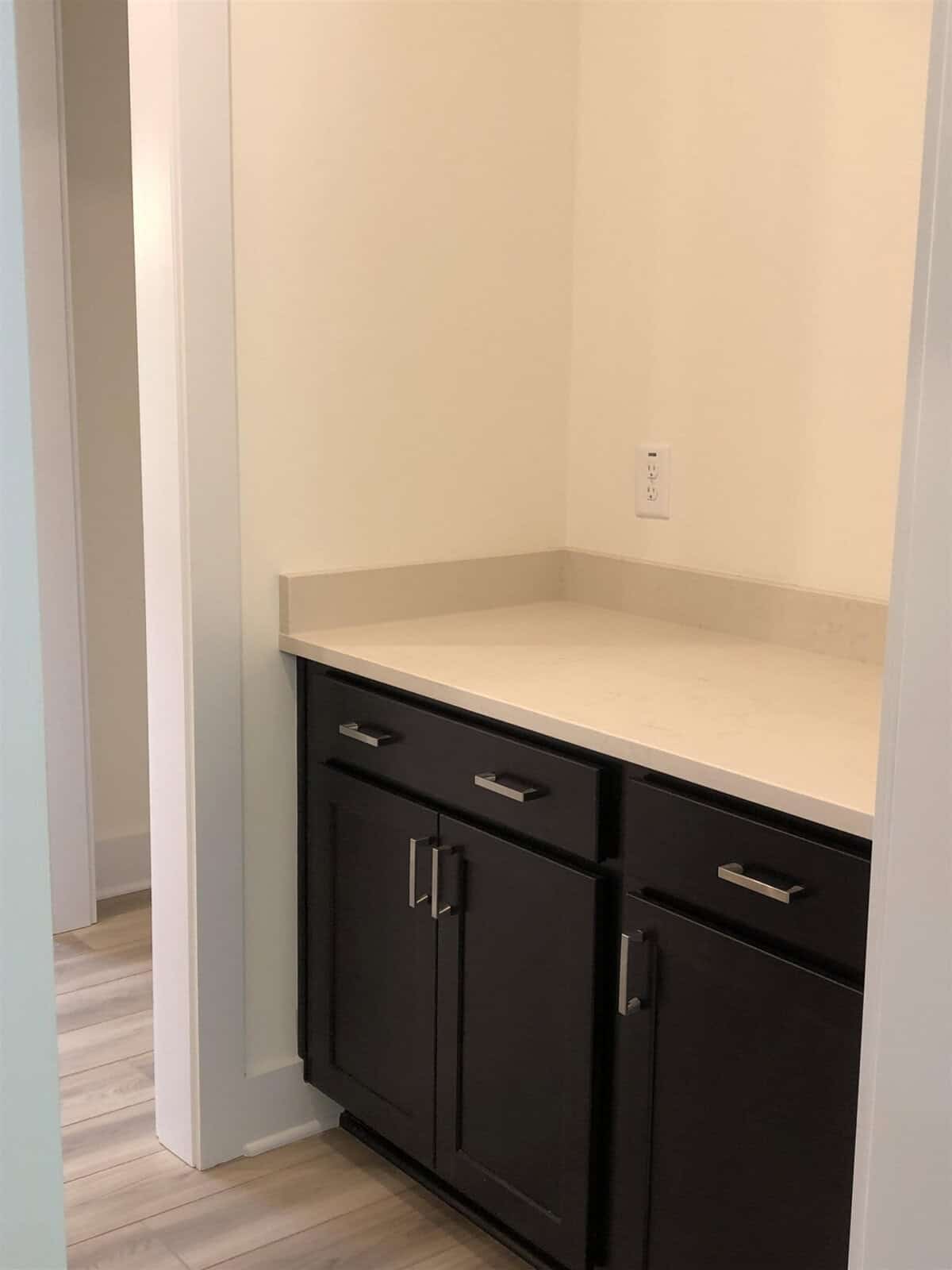 A small nook with a beige countertop, dark cabinets with silver handles, a white wall, an electrical outlet, and light-colored wood flooring. The area is well-lit and minimalistic.