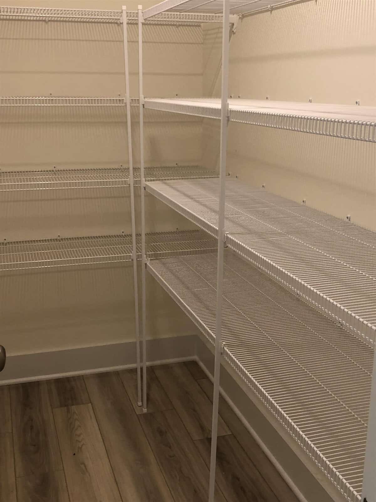 Empty walk-in pantry with white metal wire shelving on cream-colored walls. The shelves line two walls and the floor is light wood laminate. No items are present on the shelves or floor. Door handle visible on the left.