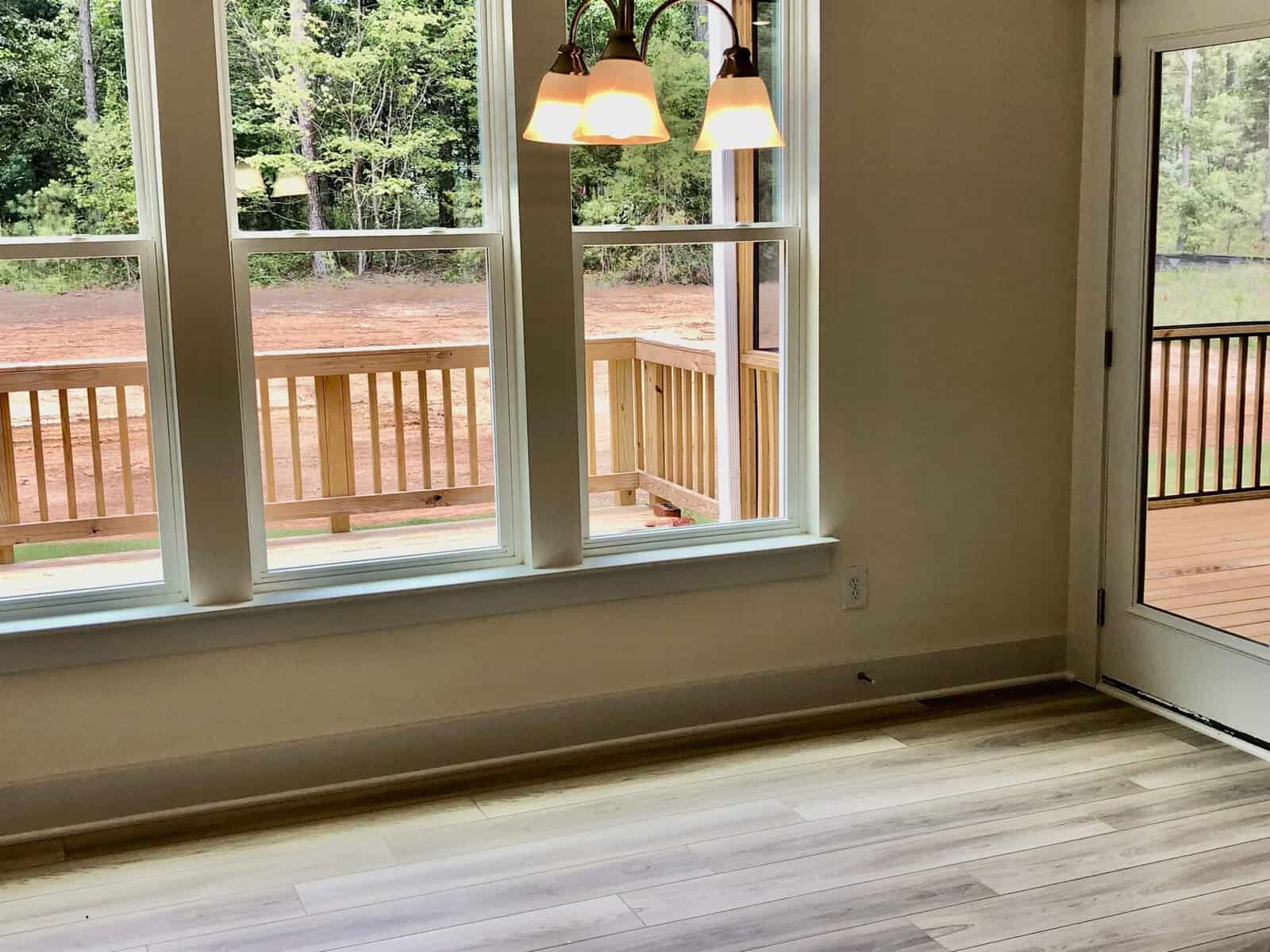 A room with light wood flooring, large windows, and a door leads to a wooden deck. The deck overlooks a dirt yard with trees in the background. Three pendant lights hang from the ceiling.