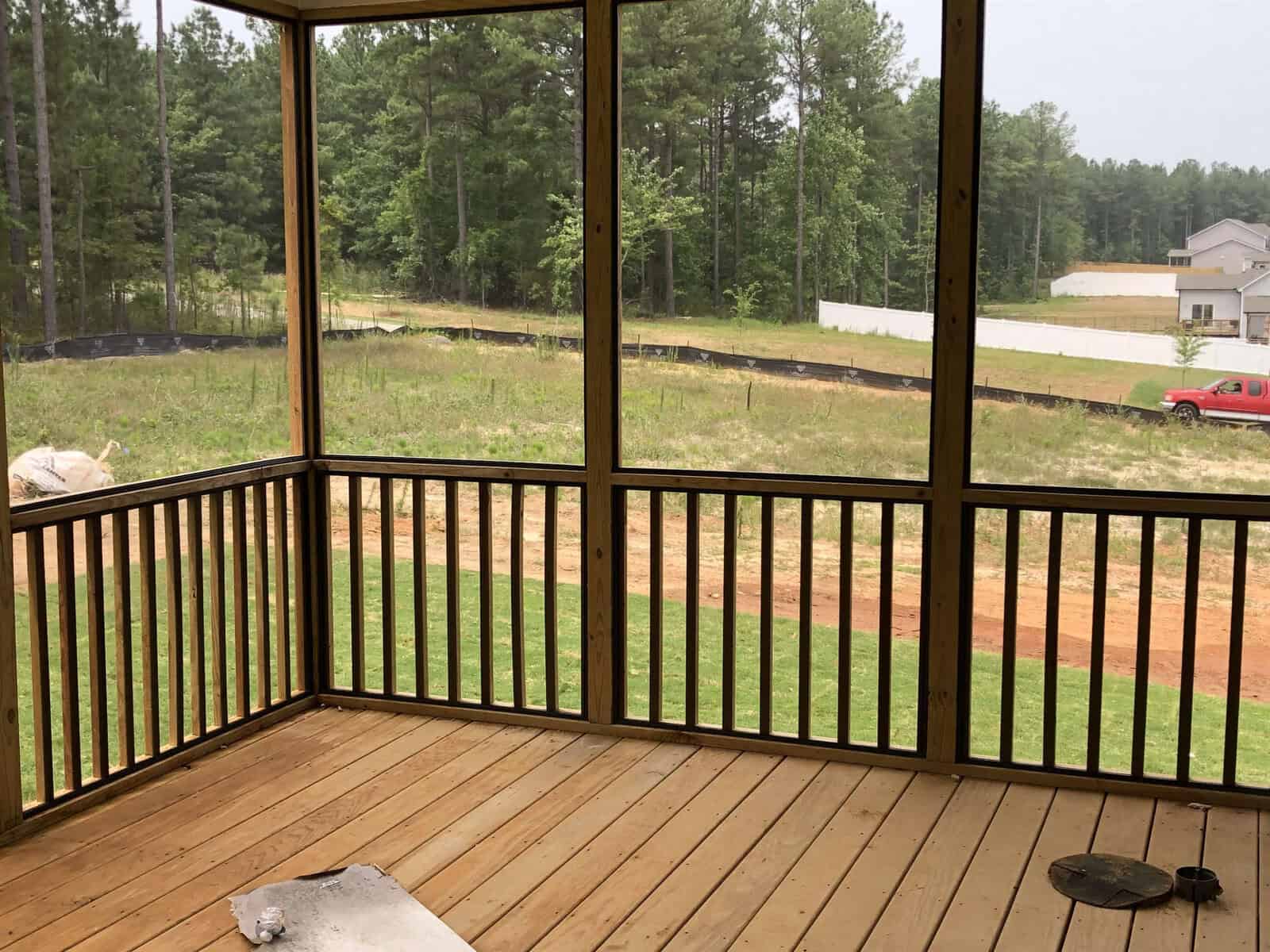 A wooden screened-in porch overlooks a grassy backyard and dirt road, with trees and a red truck visible in the background. A rolled-up mat and circular object lie on the porch floor.