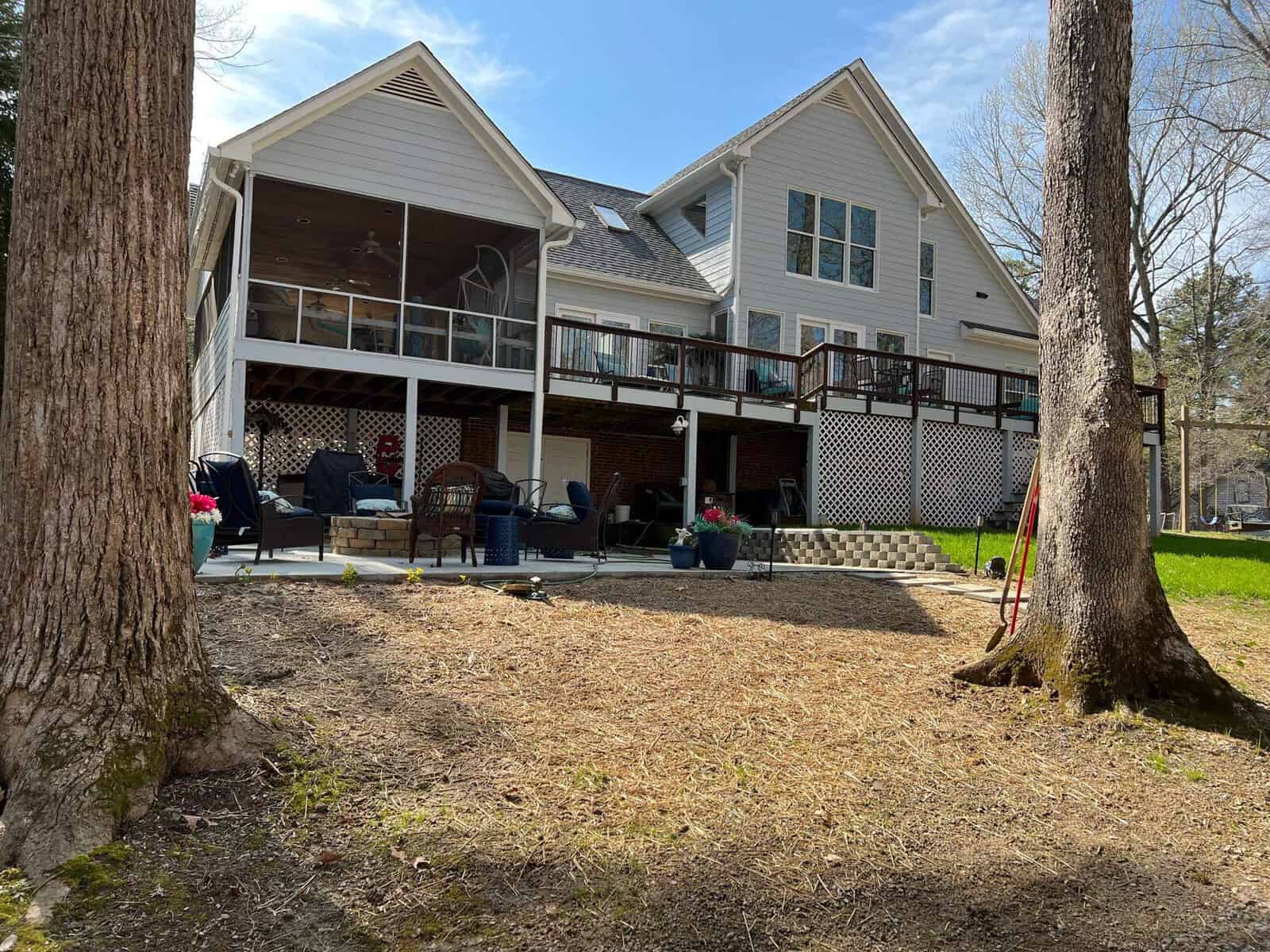 A two-story house with a large deck and outdoor seating areas surrounded by trees on a sunny day.
