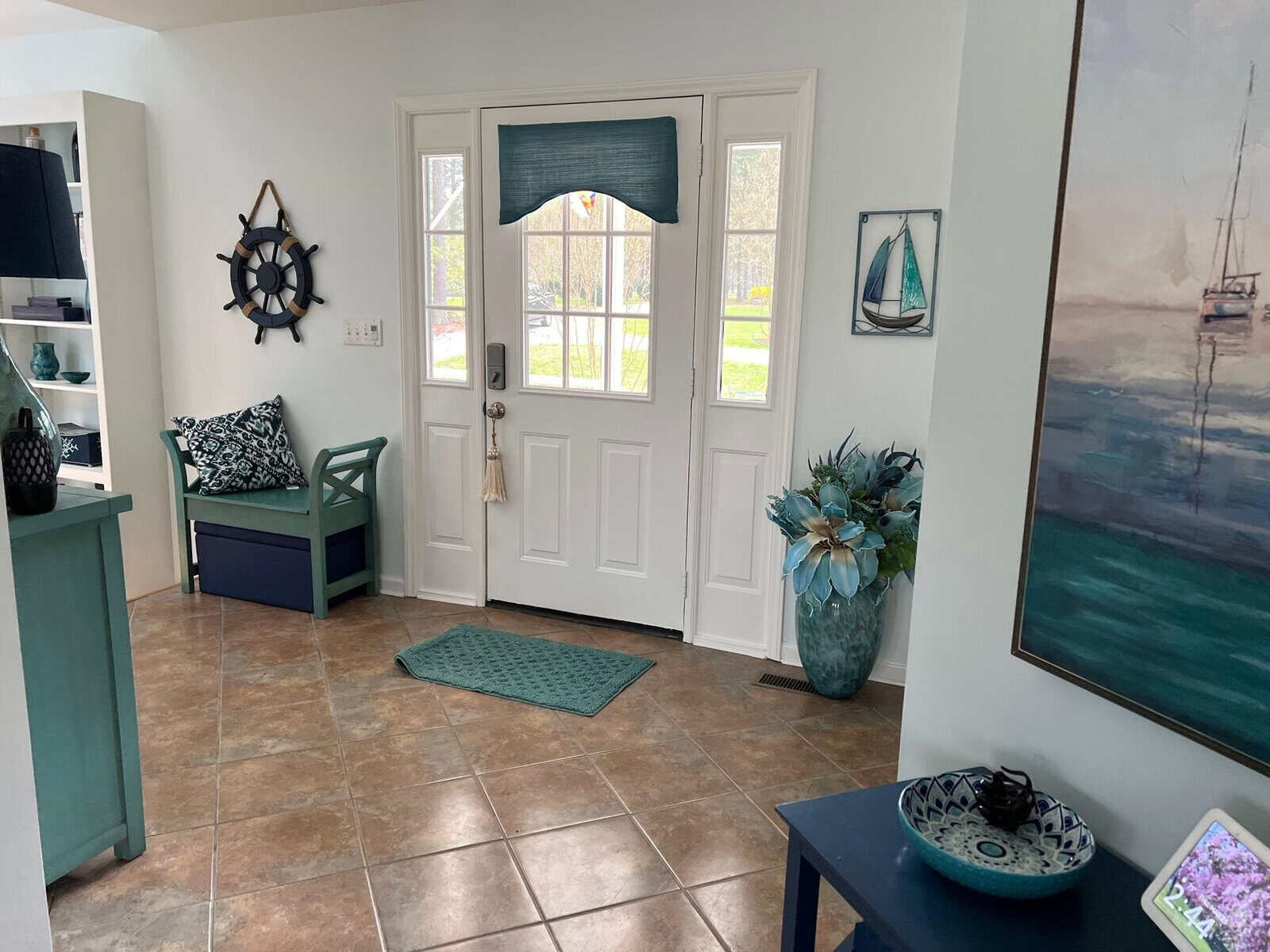 An entryway of a house with a nautical theme, featuring a white double door with a window and a blue window treatment above, a wall-mounted ship's wheel, and coastal décor including a blue bench, a green cushioned chair, and ocean-inspired artwork.