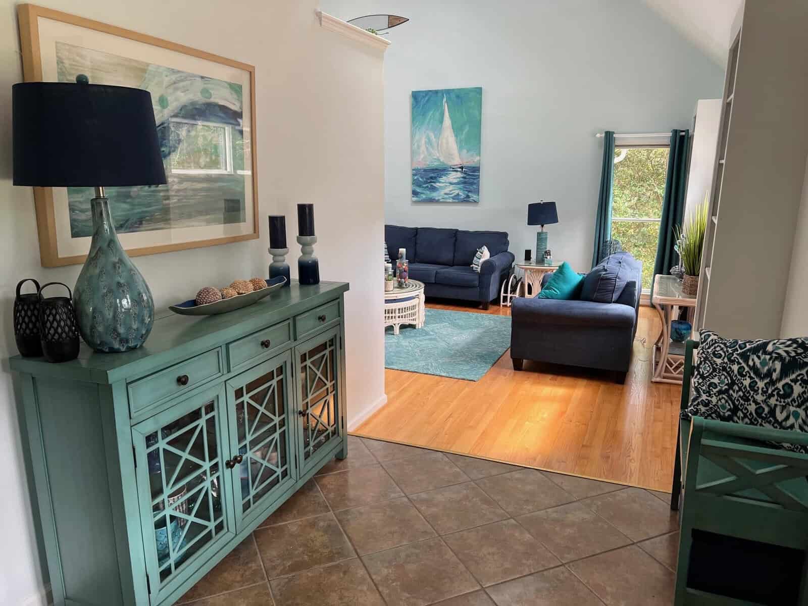 A cozy living room interior with a blue sofa, matching decorations, and paintings of maritime scenes, seen from an adjoining room with a teal cabinet and decorative items.