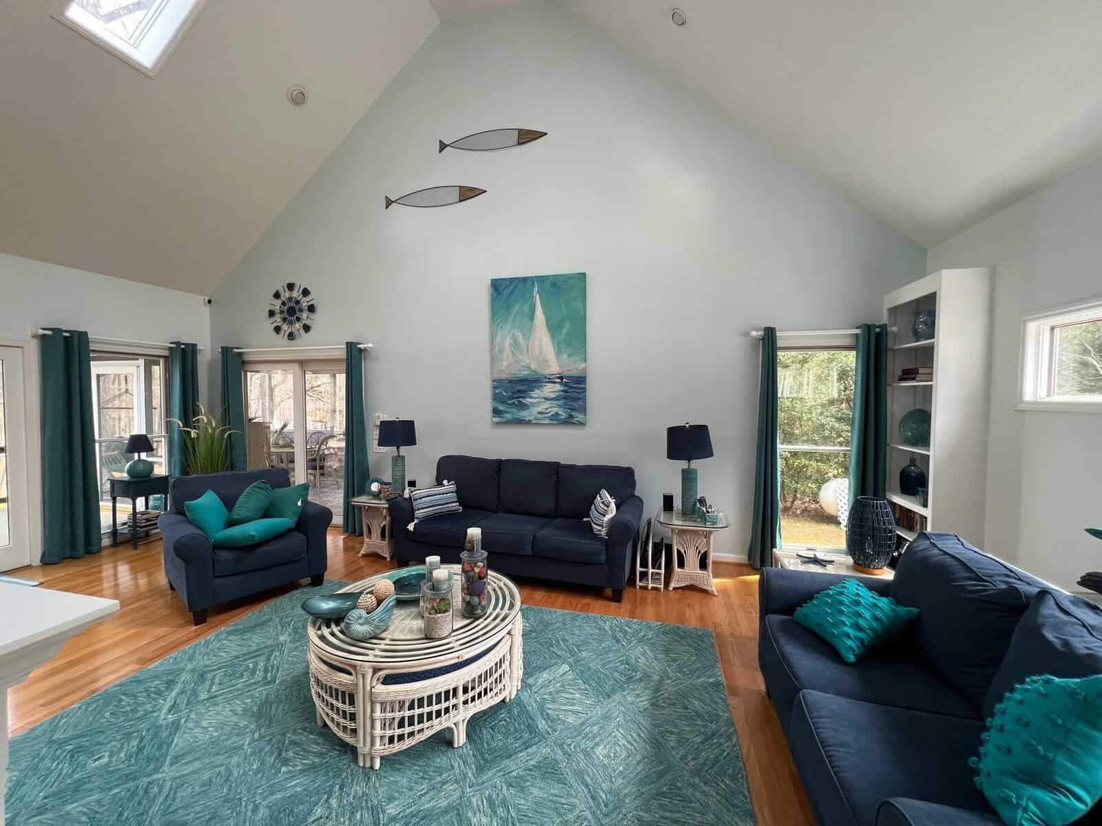 A spacious living room with vaulted ceilings, featuring a nautical theme with ocean artwork and decorative fish, two dark blue couches, a round ottoman coffee table, and multiple windows allowing for natural light.