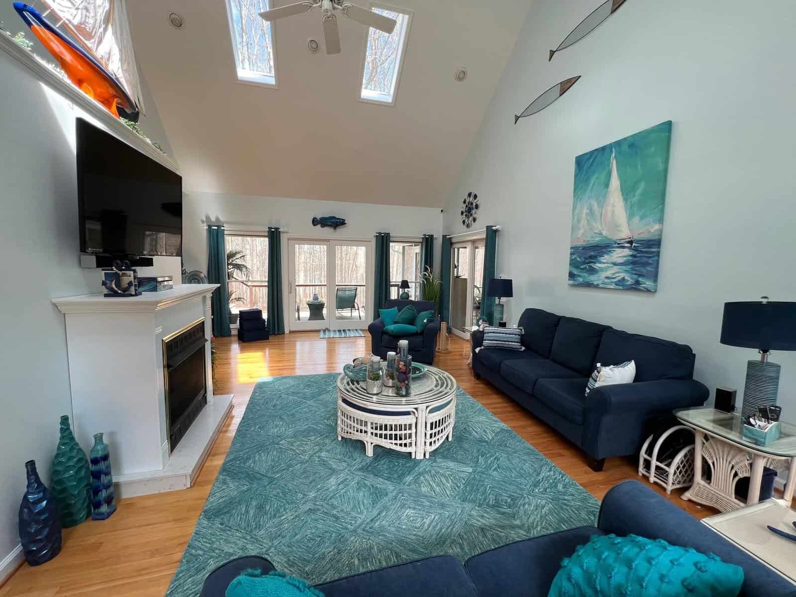 A spacious living room with high ceilings, skylights, and a seafoam green and navy blue color scheme, featuring a fireplace, large windows, and comfortable seating arrangements.