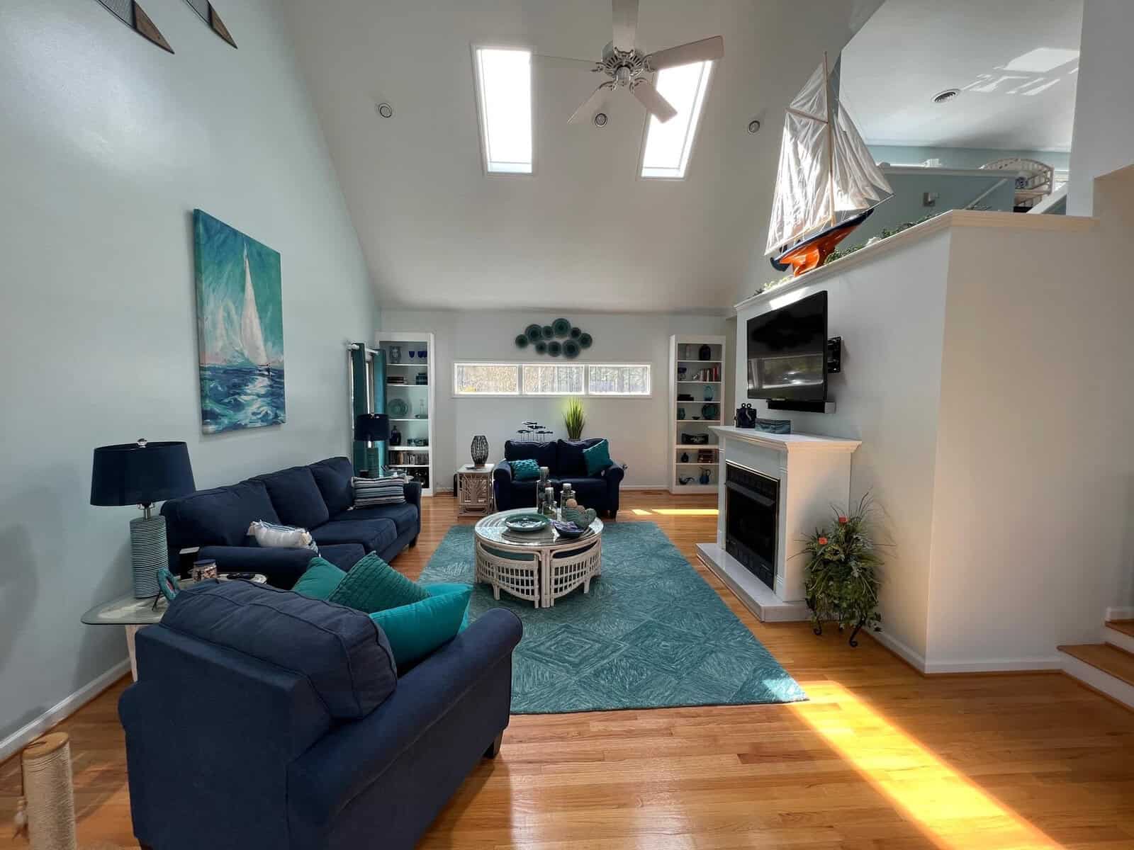 A brightly lit living room featuring a blue sofa set, a white fireplace, wooden flooring, and a vaulted ceiling with skylights.