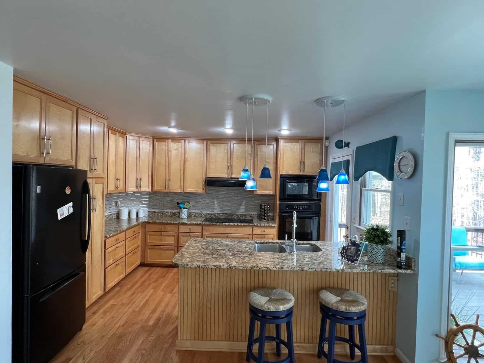 A well-lit kitchen with wooden cabinets, stainless steel appliances, a granite countertop island with bar stools, and blue pendant lights.