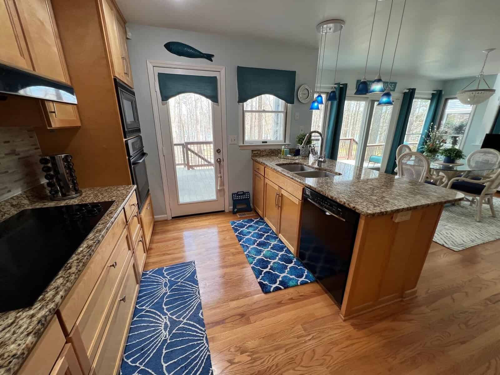 A modern kitchen with wooden cabinetry, granite countertops, and stainless steel appliances, featuring two pendant lights over the island and a view of an adjoining dining area with ample natural light.
