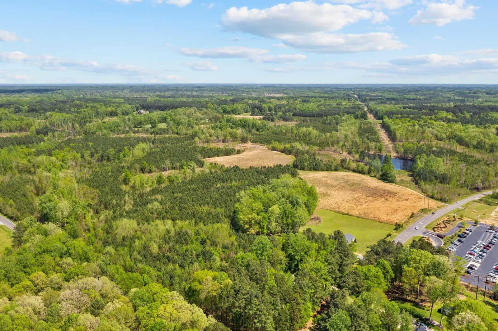 Aerial view of a vast green landscape near 00 Salem Road, with dense forests, patches of brown fields, and a winding road. A parking lot with some cars is visible in the lower right corner. The sky above is blue with fluffy white clouds hovering over Oxford's serene landscape.