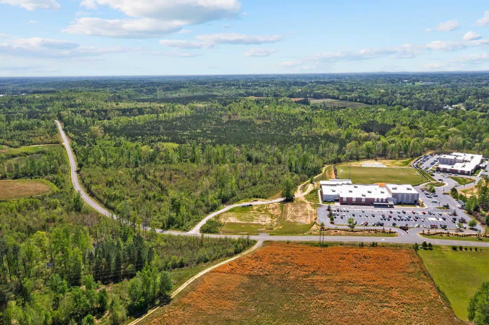 Aerial view of a rural landscape with forests, a winding road, and two buildings surrounded by parking lots. In the foreground, there are open fields. The sky is clear with scattered clouds.