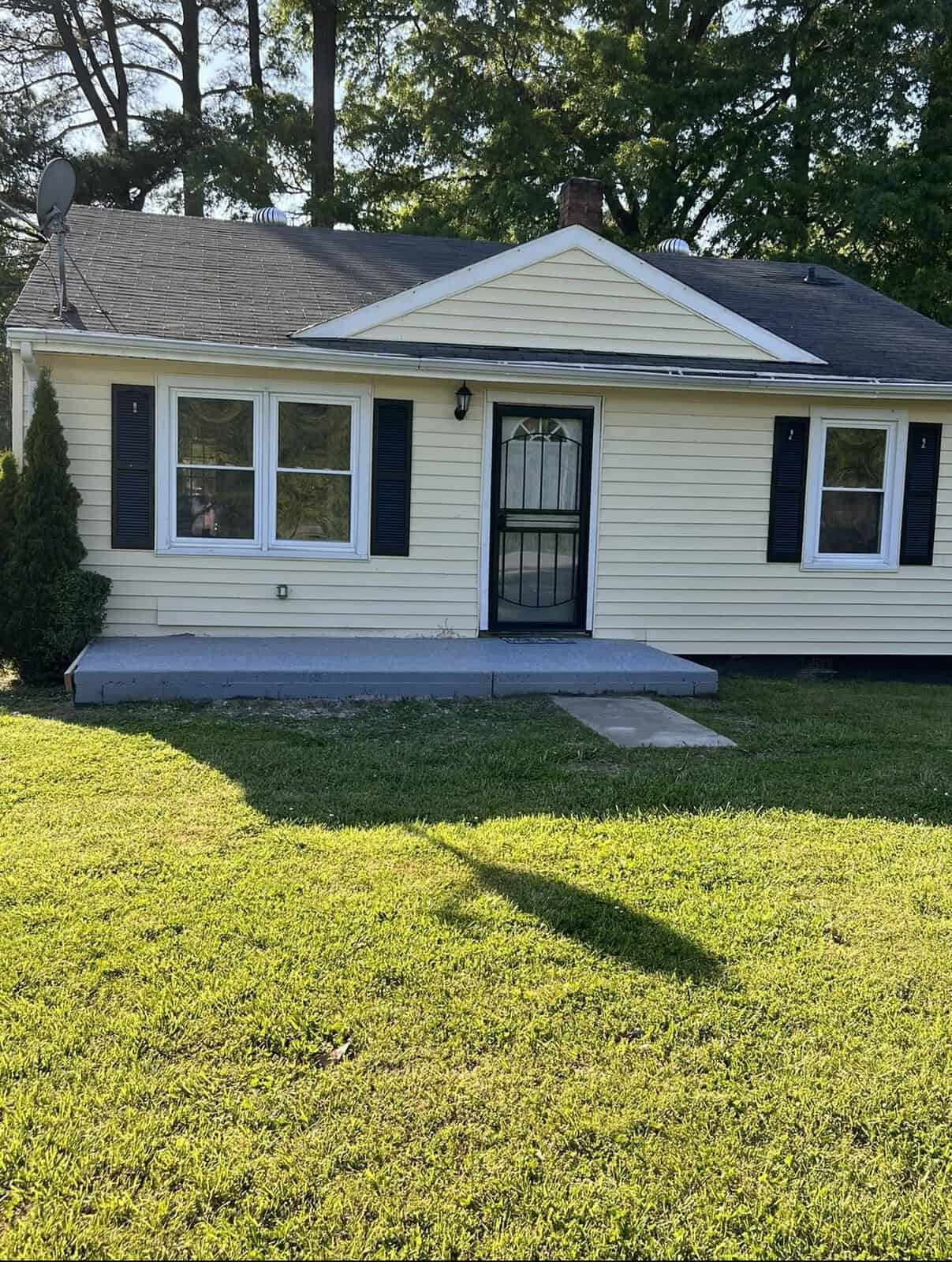 A single-story house with beige siding and white trim, featuring large front windows and a black storm door, with a well-maintained lawn in the foreground and a satellite dish on the left side.