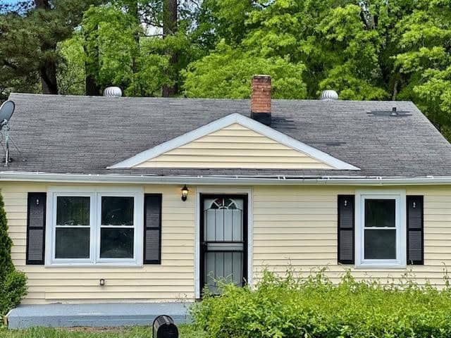 A single-story house with a yellow siding exterior, black shutters on the windows, a shingled roof with a brick chimney, and satellite dish attached.