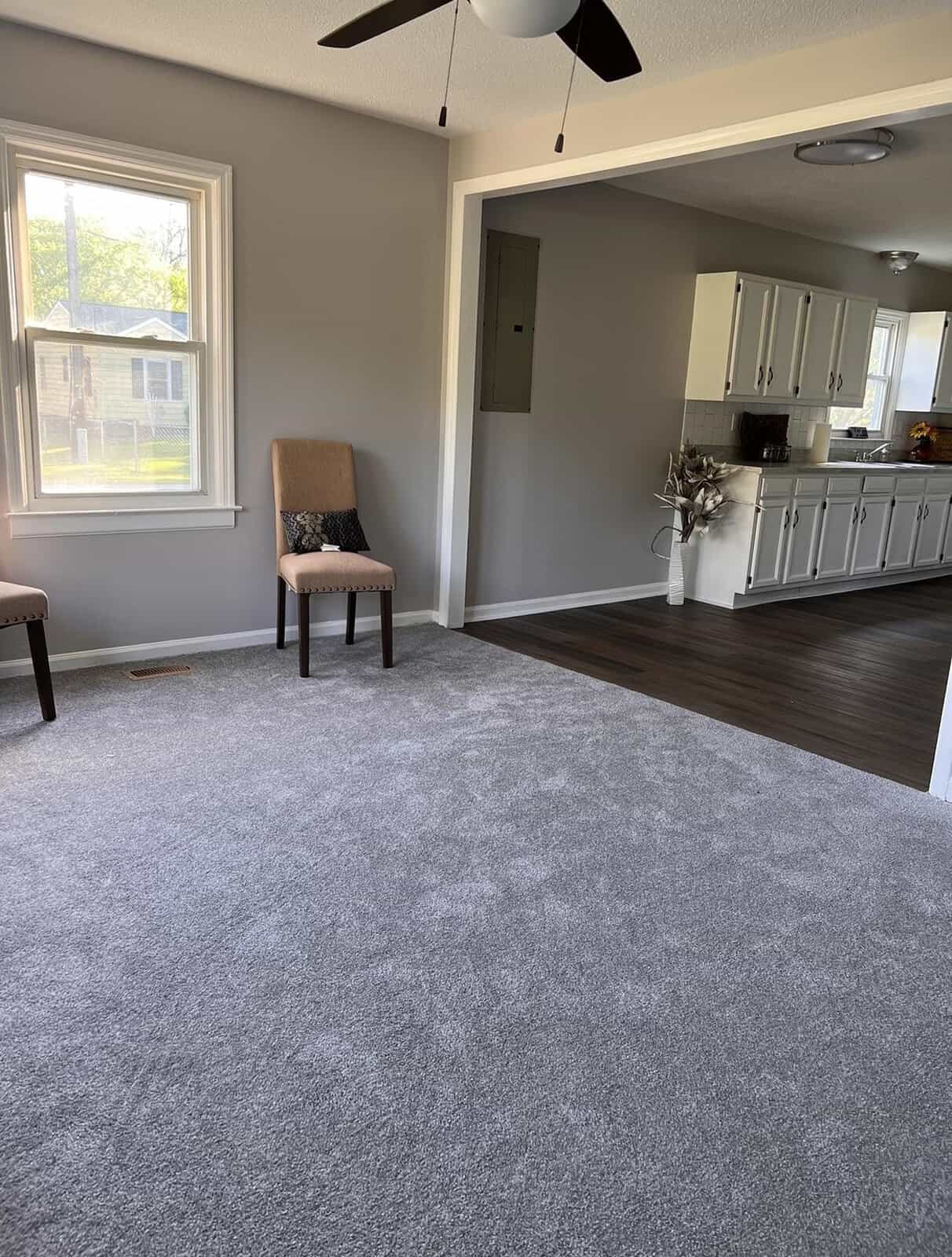 A well-lit, empty room with gray carpeting, featuring a single upholstered chair by the window and an adjoining kitchen with white cabinets and stainless steel appliances.