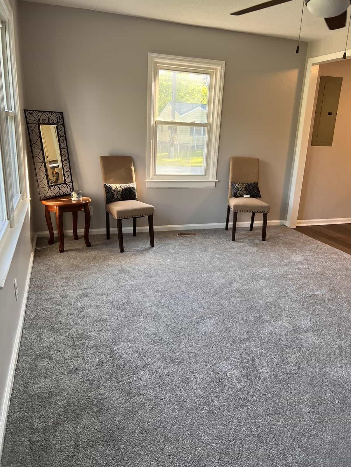 A well-lit room with a gray carpet featuring two chairs with patterned upholstery near a window, a side table with a decorative mirror above it, and a ceiling fan.