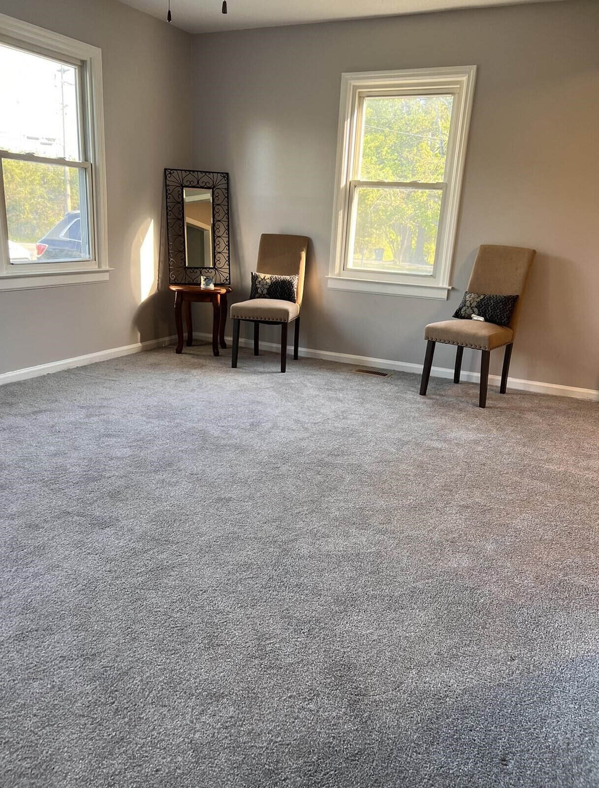 A sparsely furnished room with two chairs, each with a decorative pillow, a standing mirror, and two windows allowing in natural light.