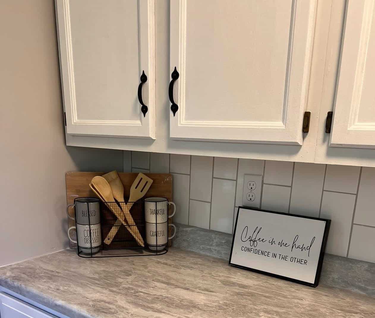 A corner of a kitchen countertop featuring a cutting board with wooden utensils, two decorated mugs, and a framed sign with the quote "coffee in one hand, confidence in the other" against a tiled backsplash and under white cabinets.