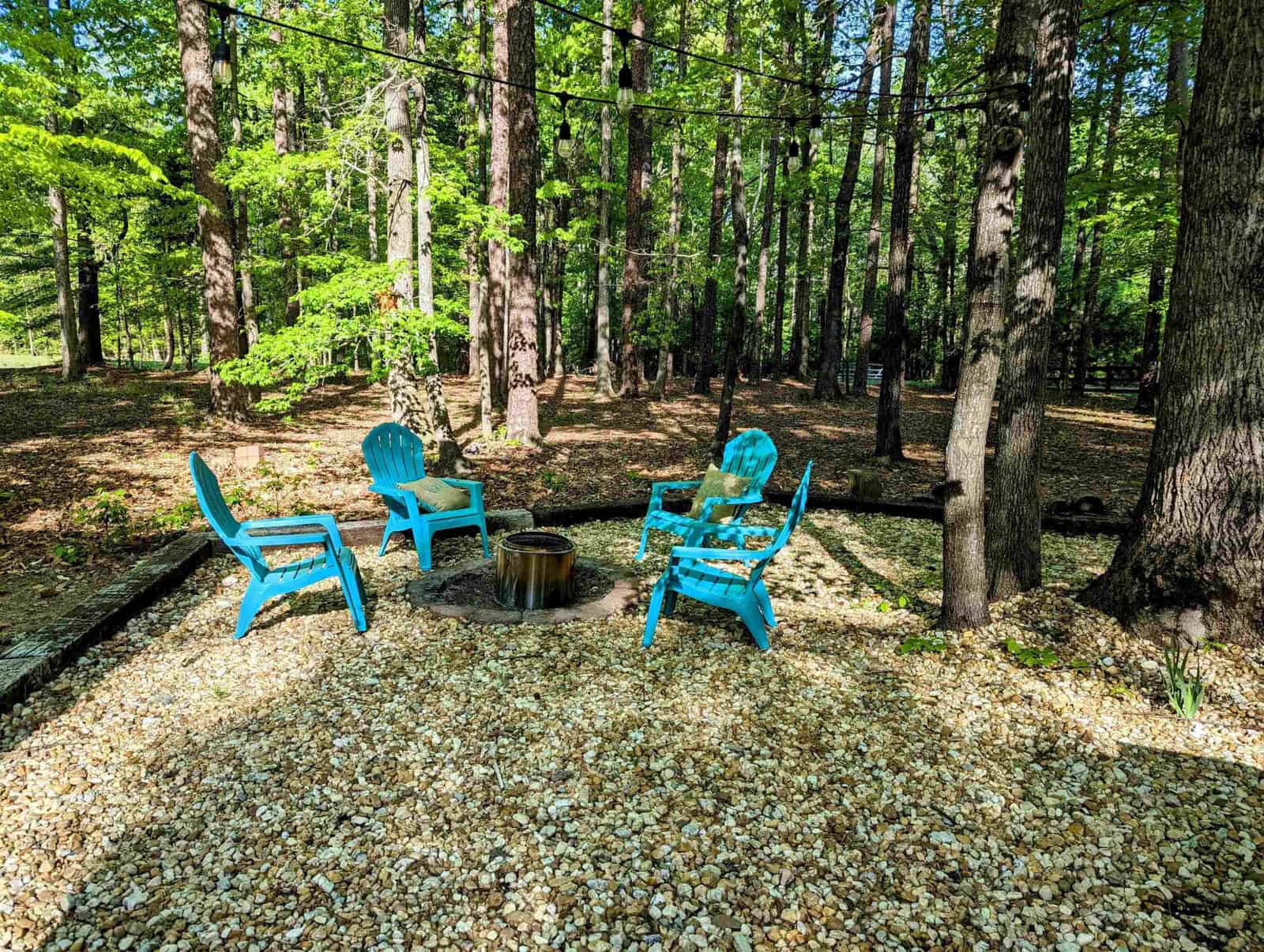 Three blue adirondack chairs arranged in a semi-circle around a small fire pit, set on a gravel surface in a forested area with sunlight filtering through the trees.